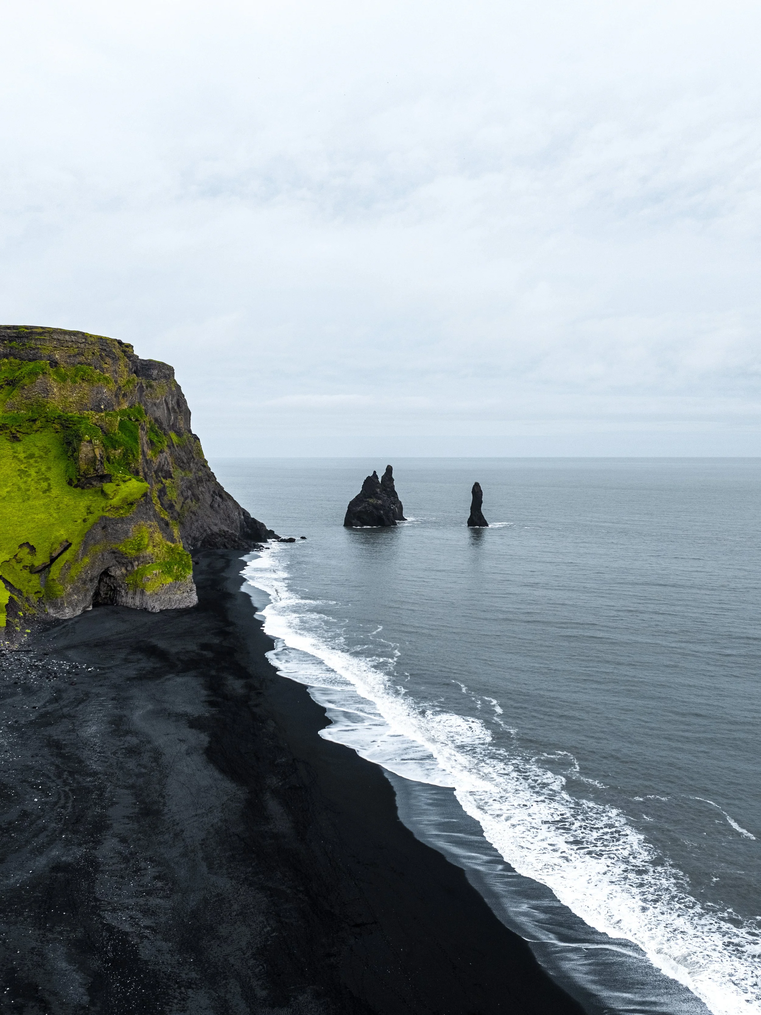 Cliffs with green grass at the top, black sand beach, and sharp volcanic rocks rising from the sea under a cloudy sky