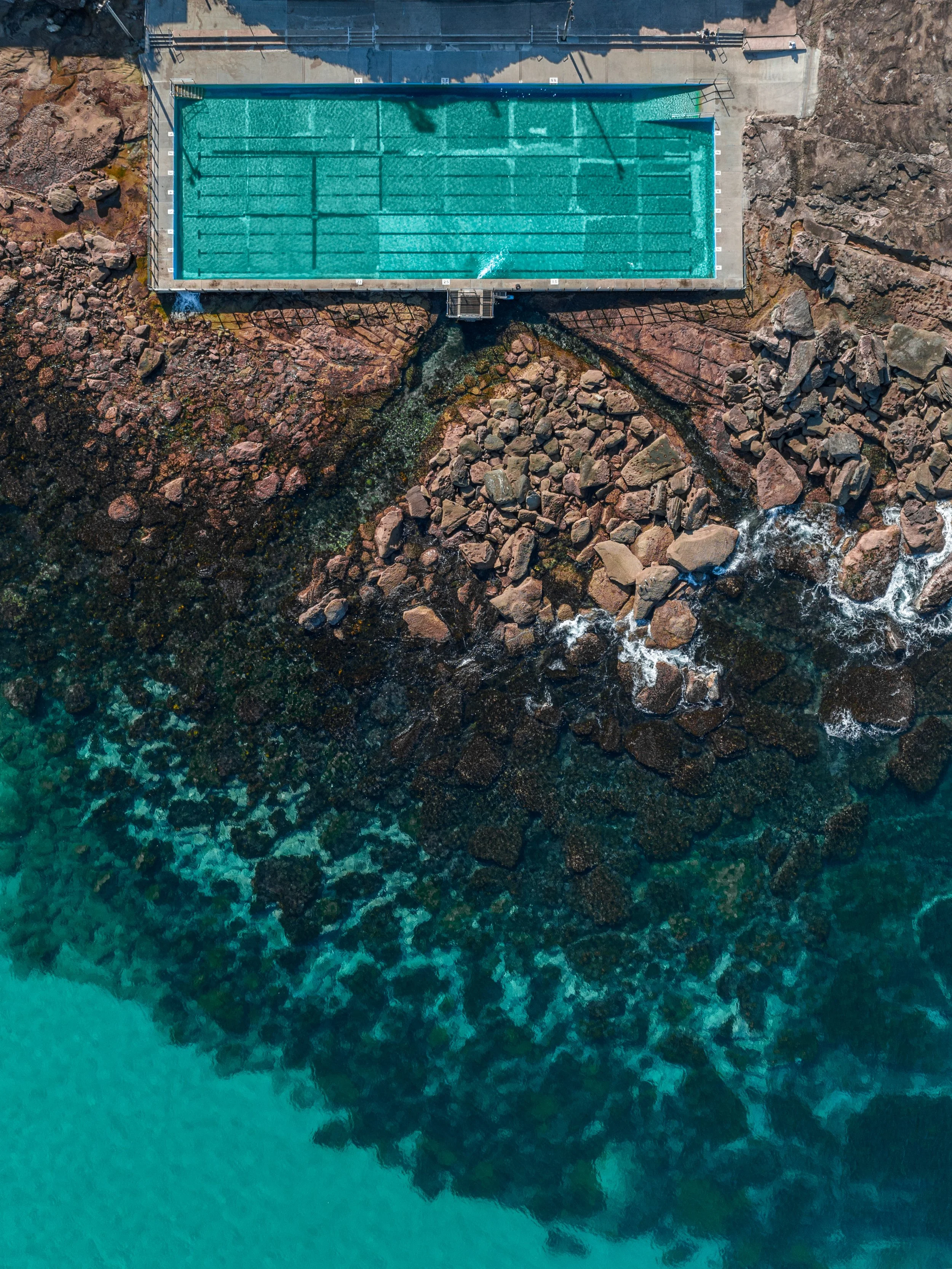 An aerial view of a rectangular outdoor swimming pool surrounded by rocky terrain and a concrete area.