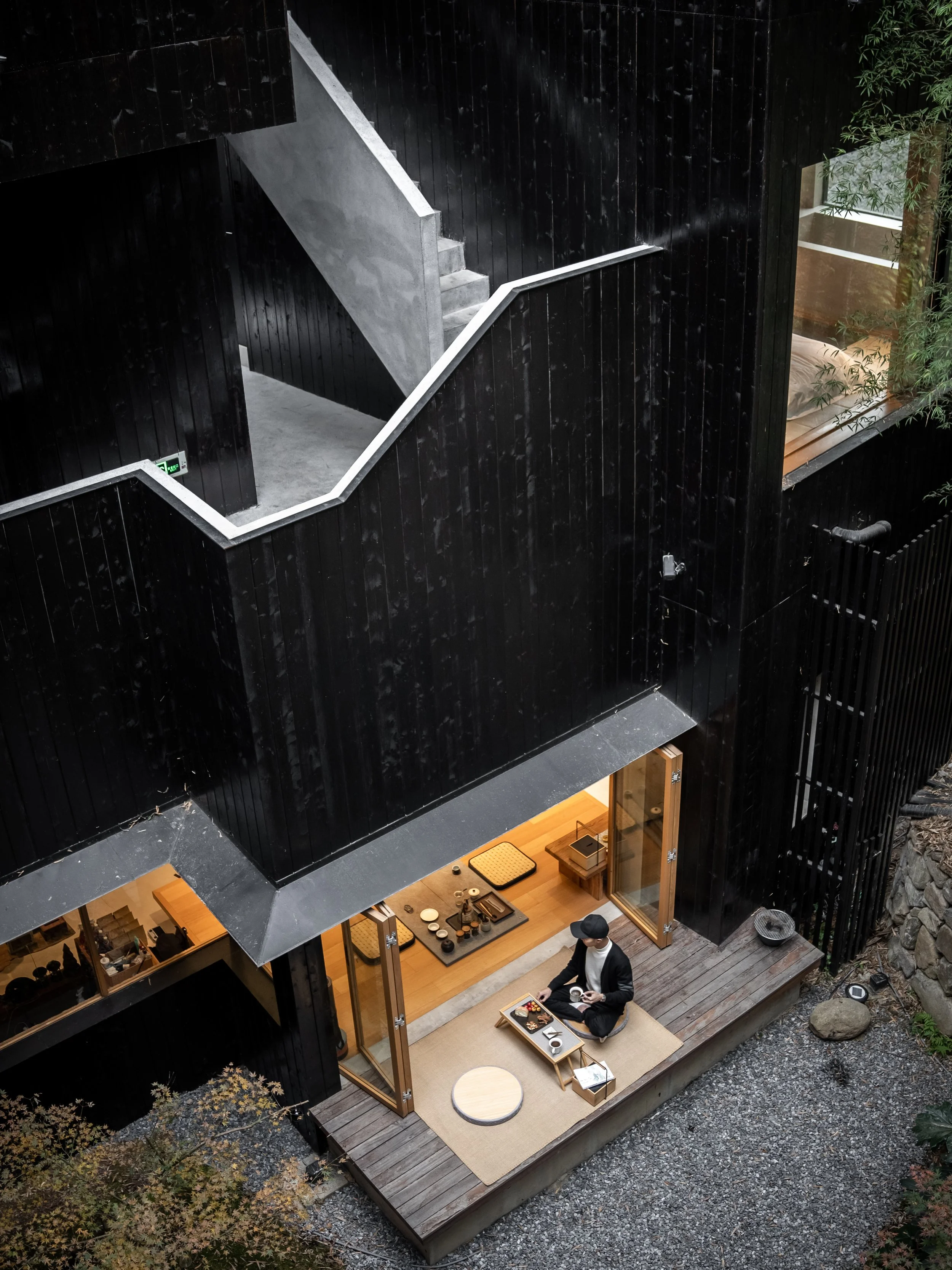 An aerial view of a modern black house with multiple levels. The lower level features a cozy outdoor patio with a person sitting on the floor, a small wooden table with objects, and a striped rug. The upper level has a staircase leading up to the sec