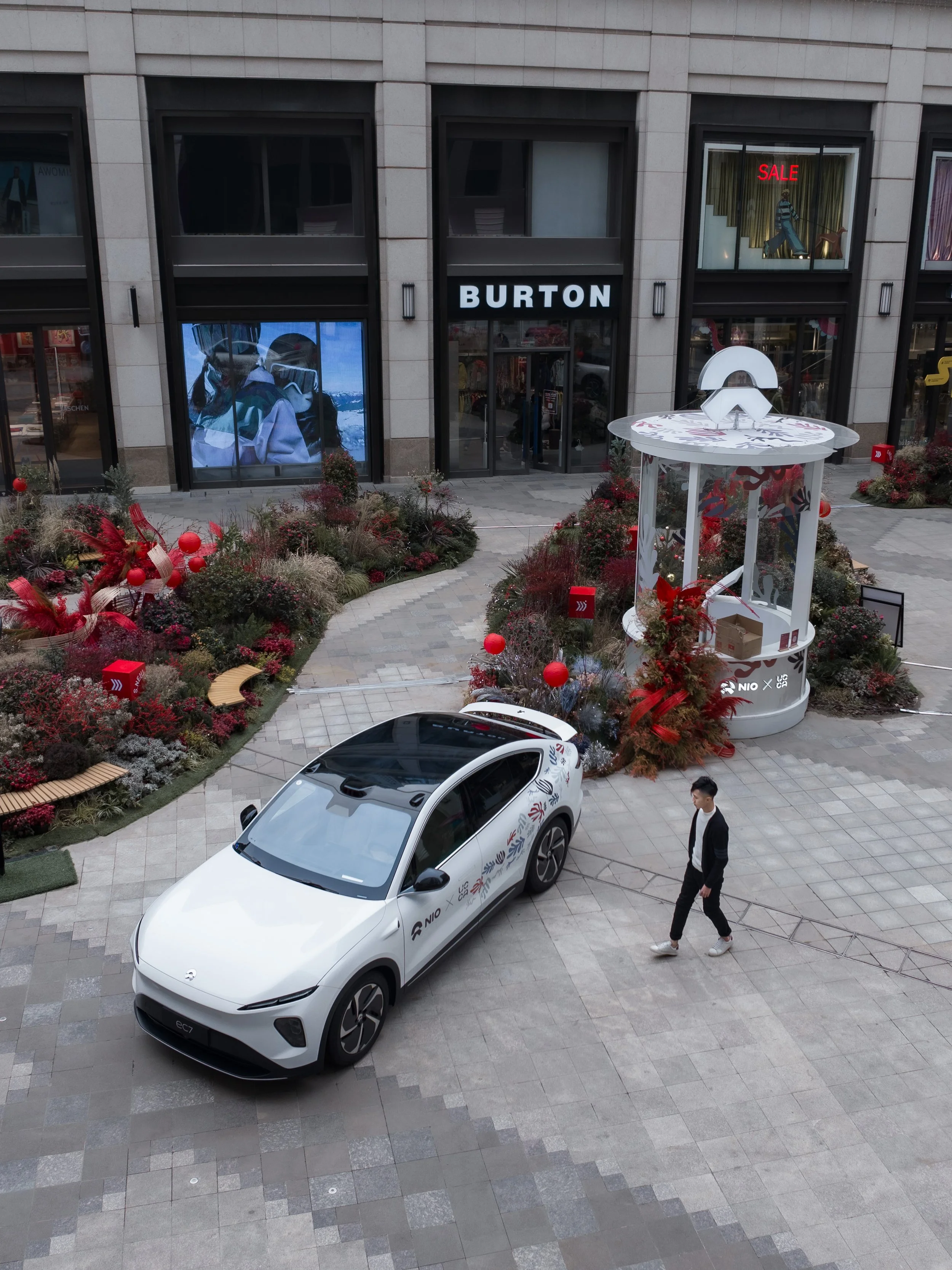 A white electric vehicle parked on a paved area in front of a store named Burton, with a man walking nearby. The store has large windows, one displaying a sale sign, and is decorated with holiday plants and ornaments.