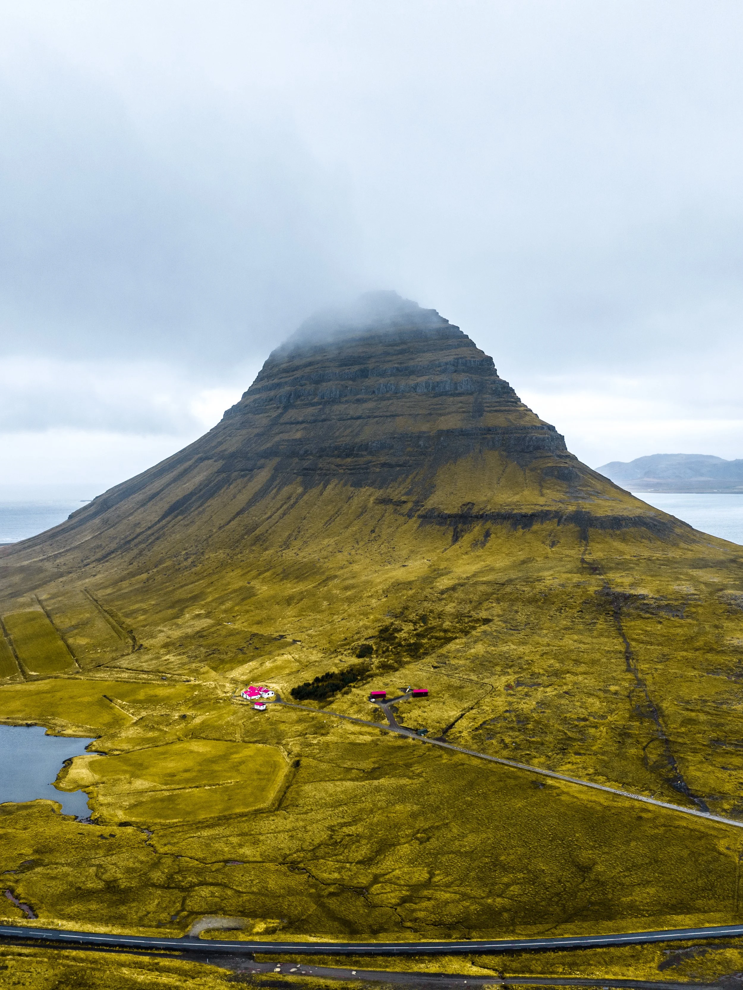 A mountain with layered rock formations, surrounded by green grass and a cloudy sky.