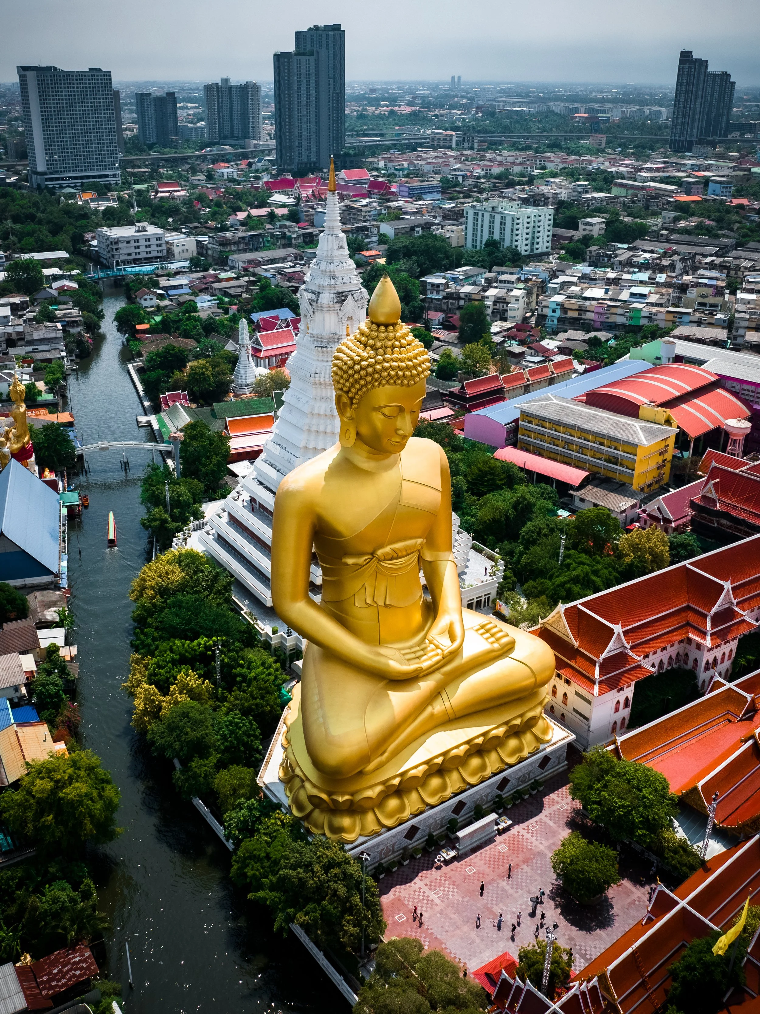 A large golden Buddha statue sitting in a meditative pose overlooking a cityscape with high-rise buildings, temples, and a river.