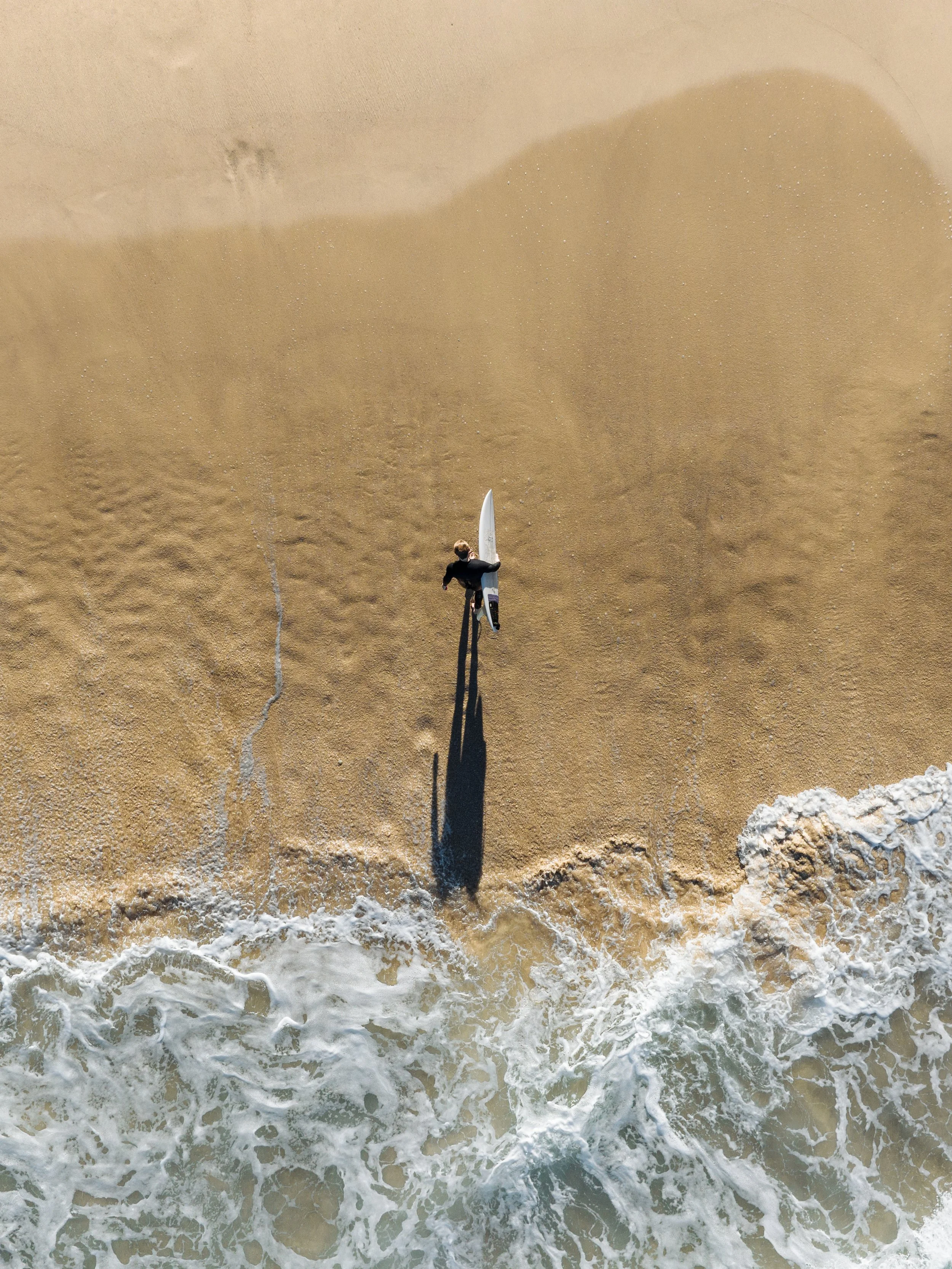 An aerial view of a person holding a surfboard walking along the sandy beach, with foamy waves washing ashore.