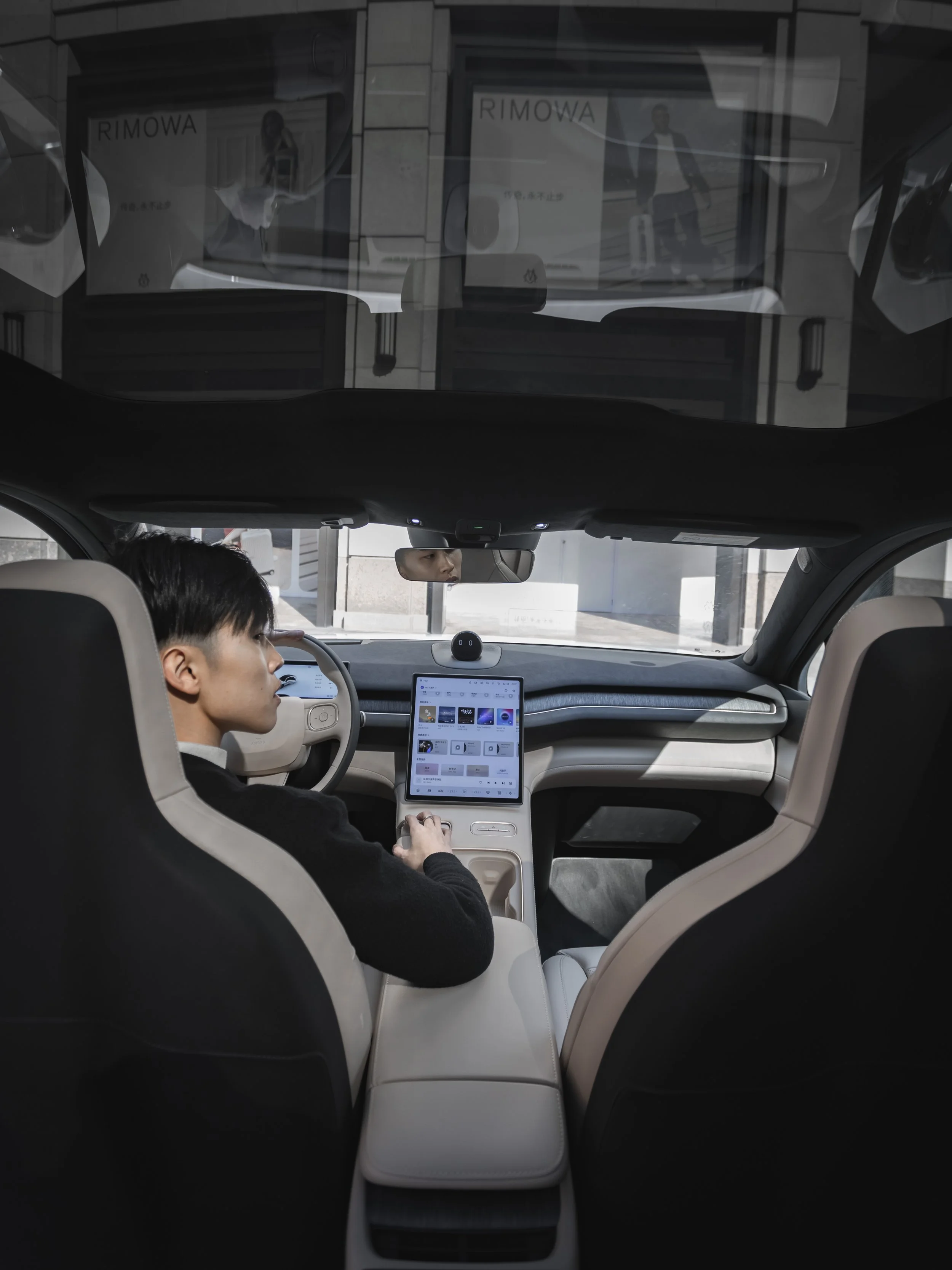 View from inside a car showing a young man sitting in the passenger seat looking at an in-car infotainment screen, with a reflection of a woman walking outside with a suitcase visible on the windshield.
