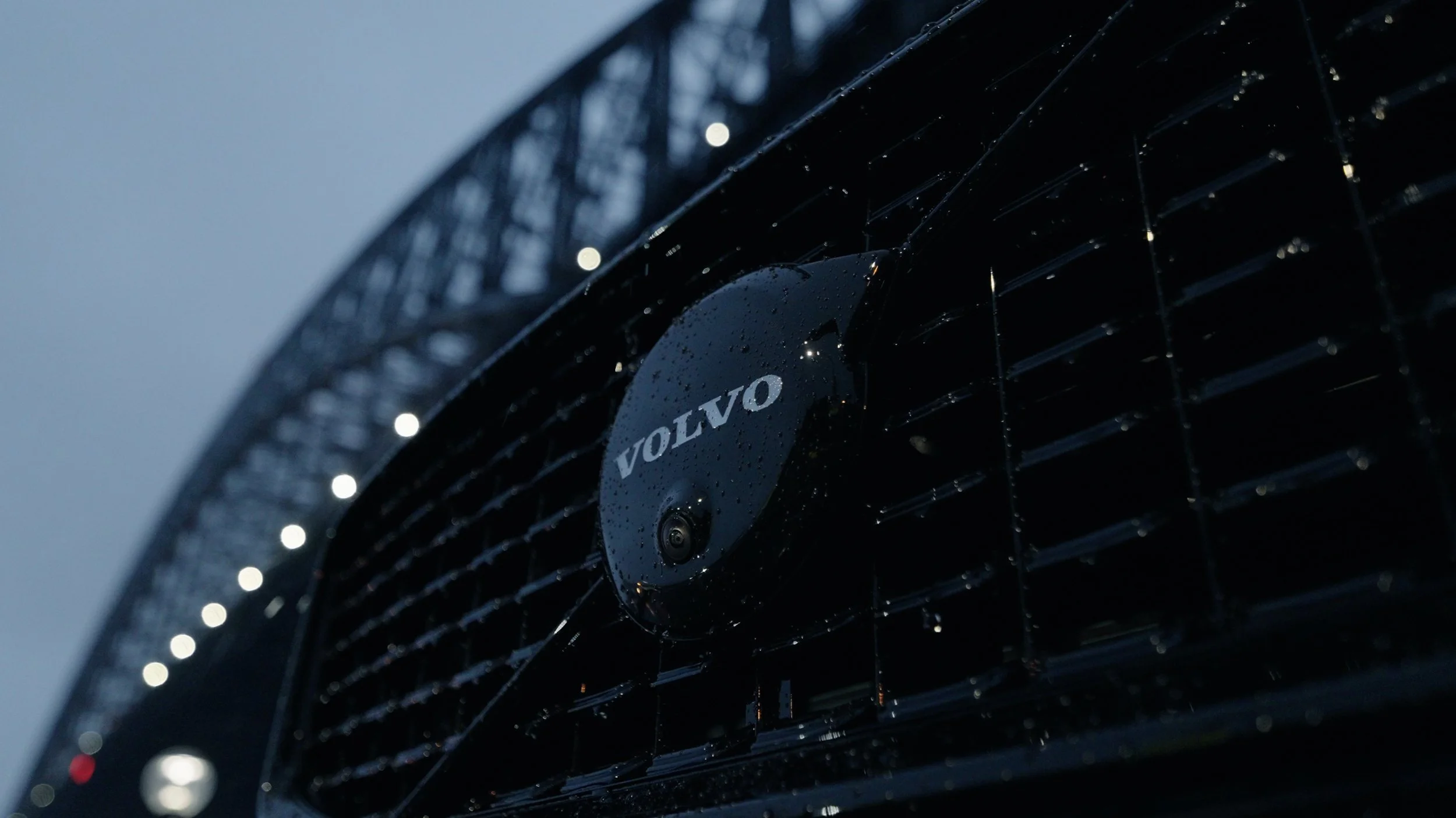 Close-up of a black Volvo car grille with raindrops, featuring the Volvo logo in the center.