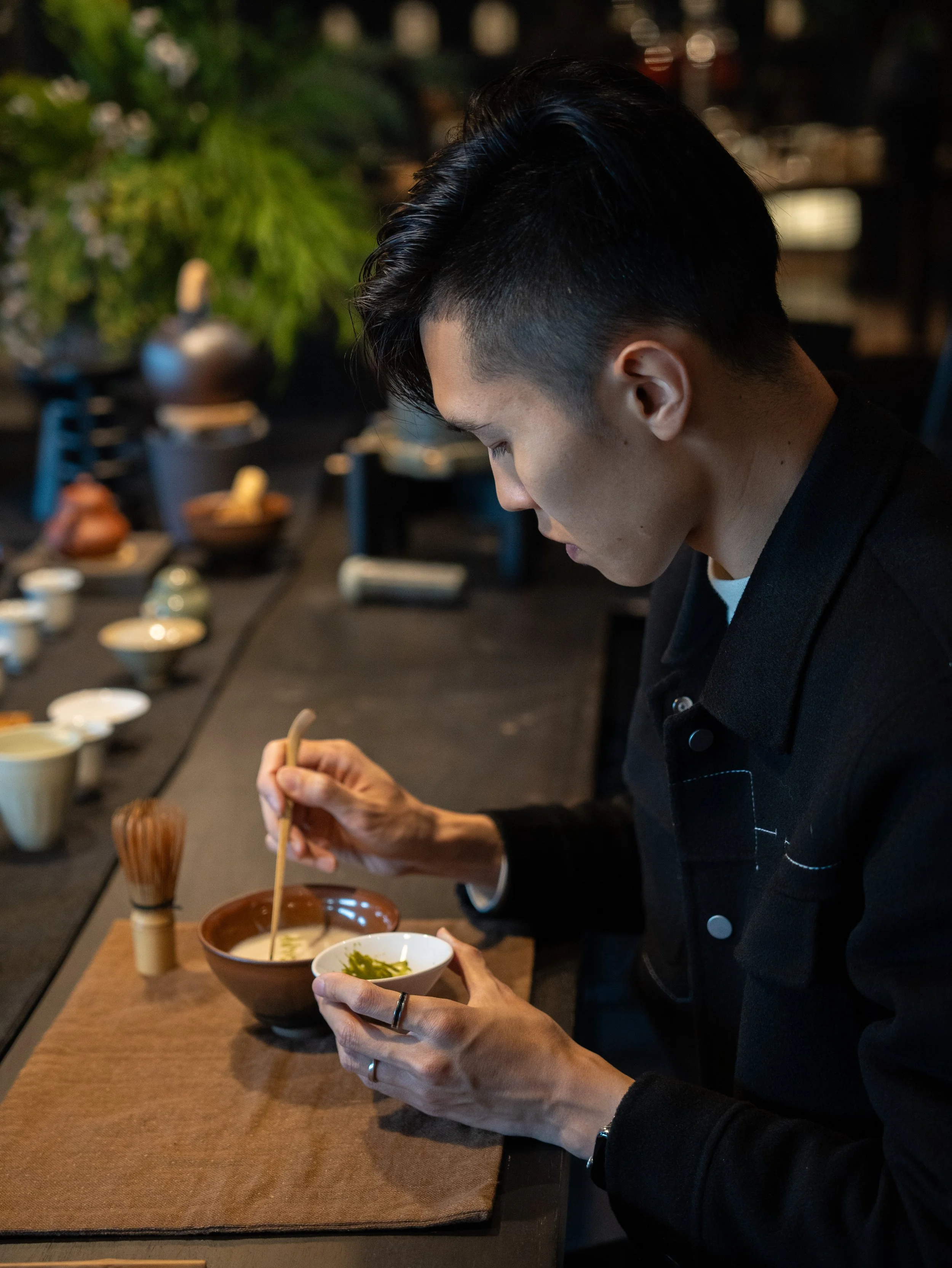A person with short black hair, wearing a black jacket, is sitting at a table in a restaurant, preparing to eat a bowl of Japanese ramen. The table has various bowls and utensils, and the background is blurred with dim lighting and green plants.