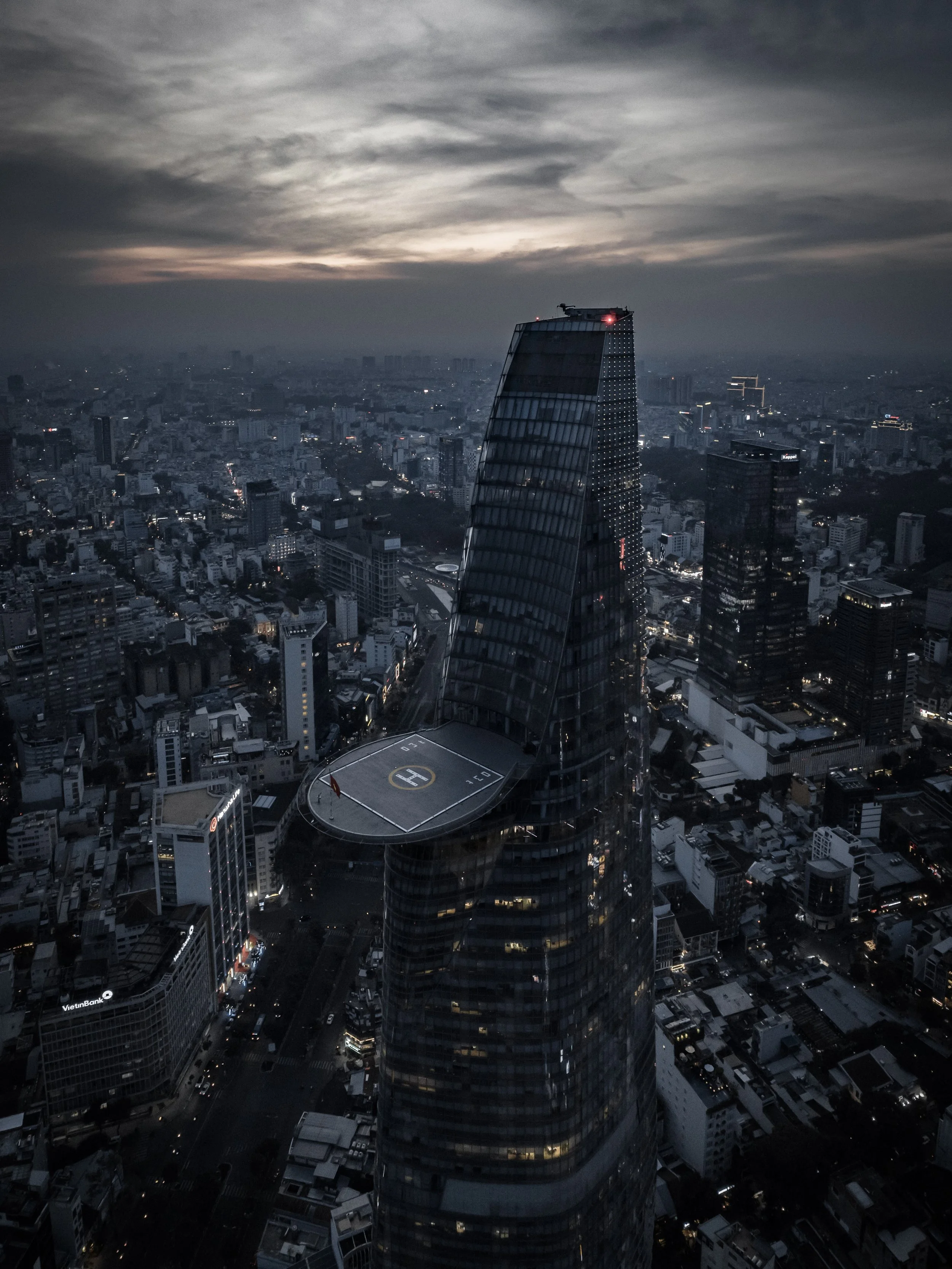 Nighttime aerial view of a city skyline featuring a tall skyscraper with a helipad on top, surrounded by numerous buildings and streets illuminated with city lights, under a cloudy sky.