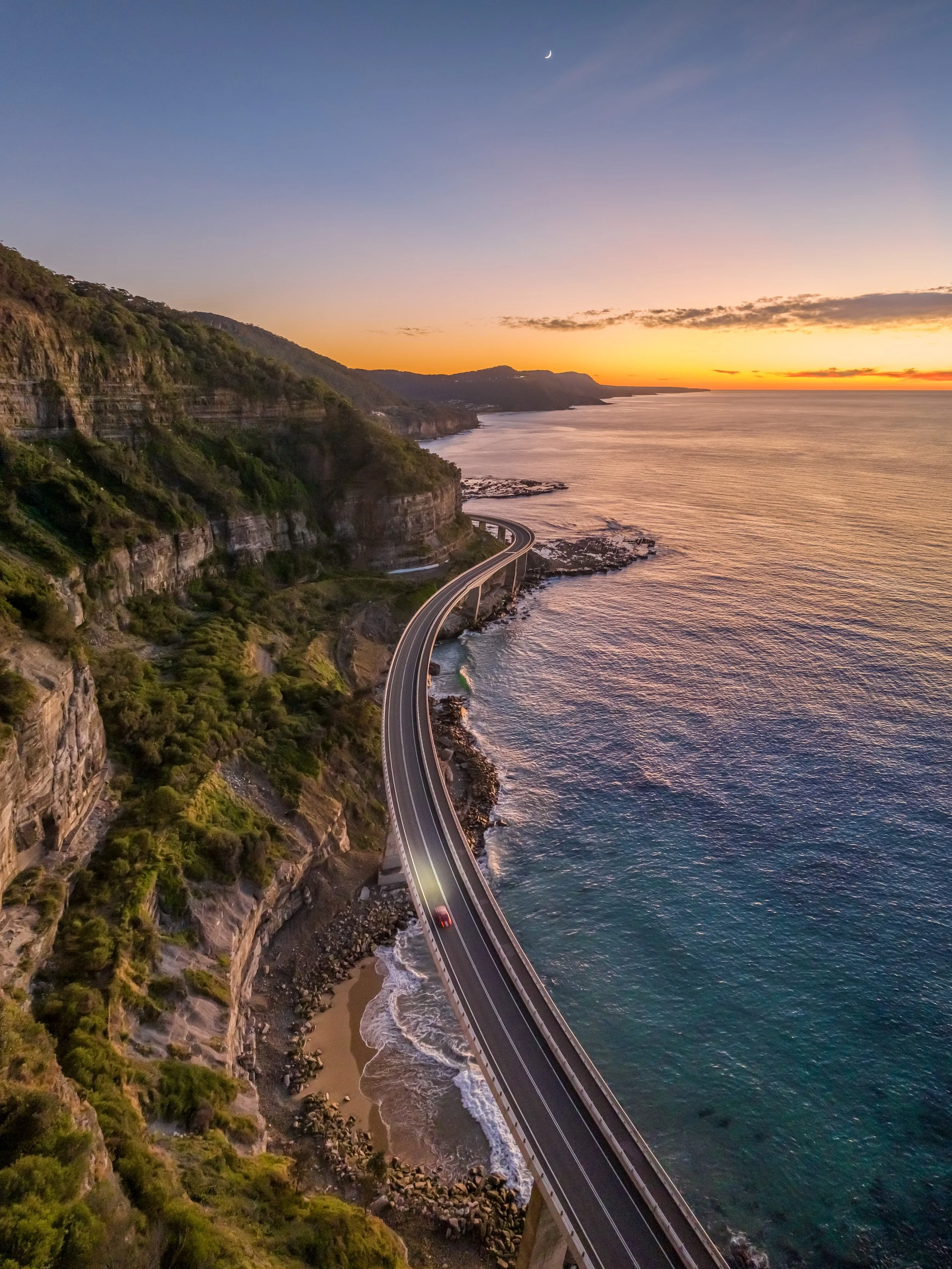 A coastal landscape at sunset with a winding road along the cliffs, the ocean on the right, and a crescent moon in the sky.