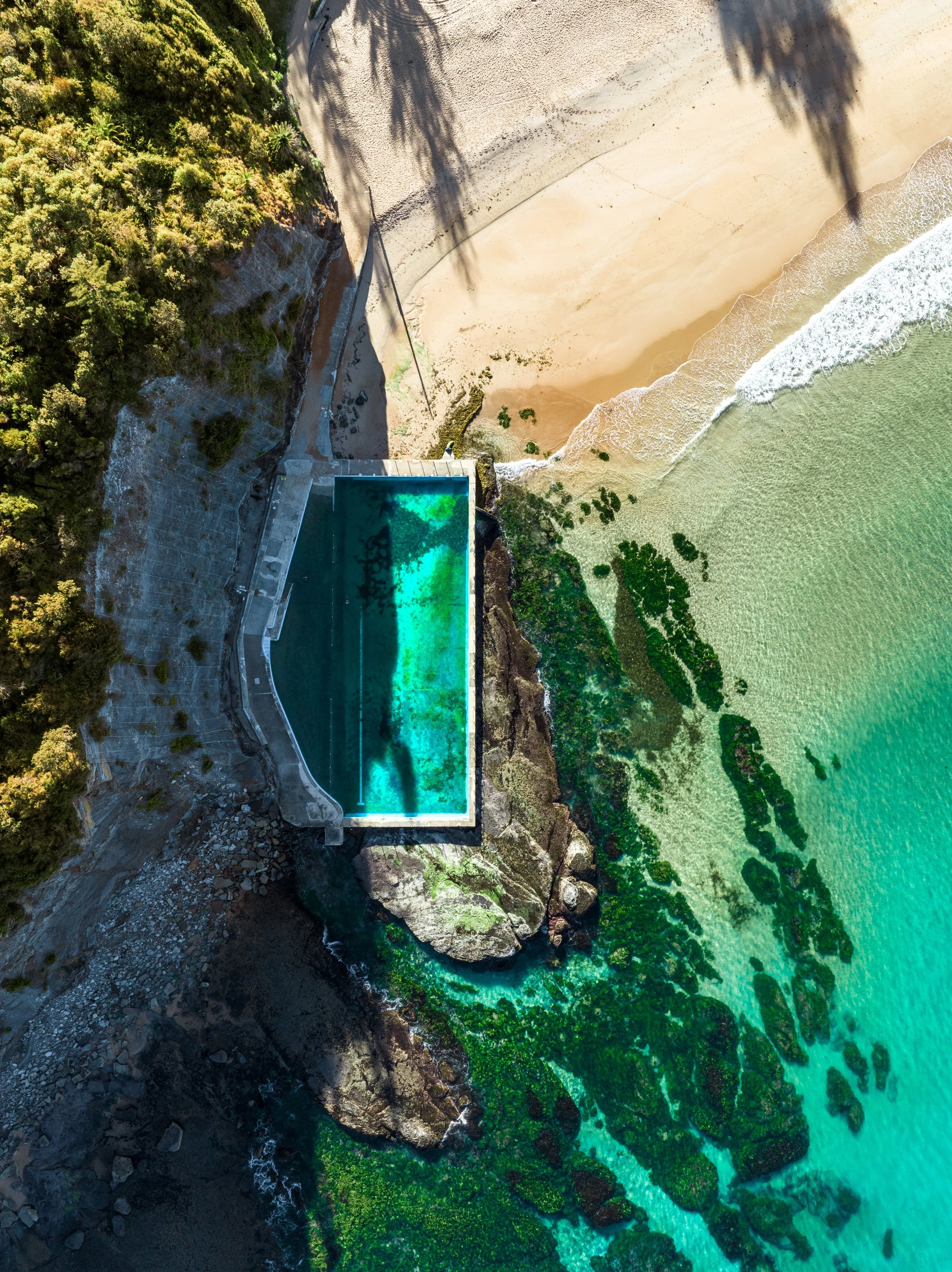 An aerial view of a beach with golden sand, turquoise water, and a freshwater swimming pool built into the rocky coastline, surrounded by green trees and plants.
