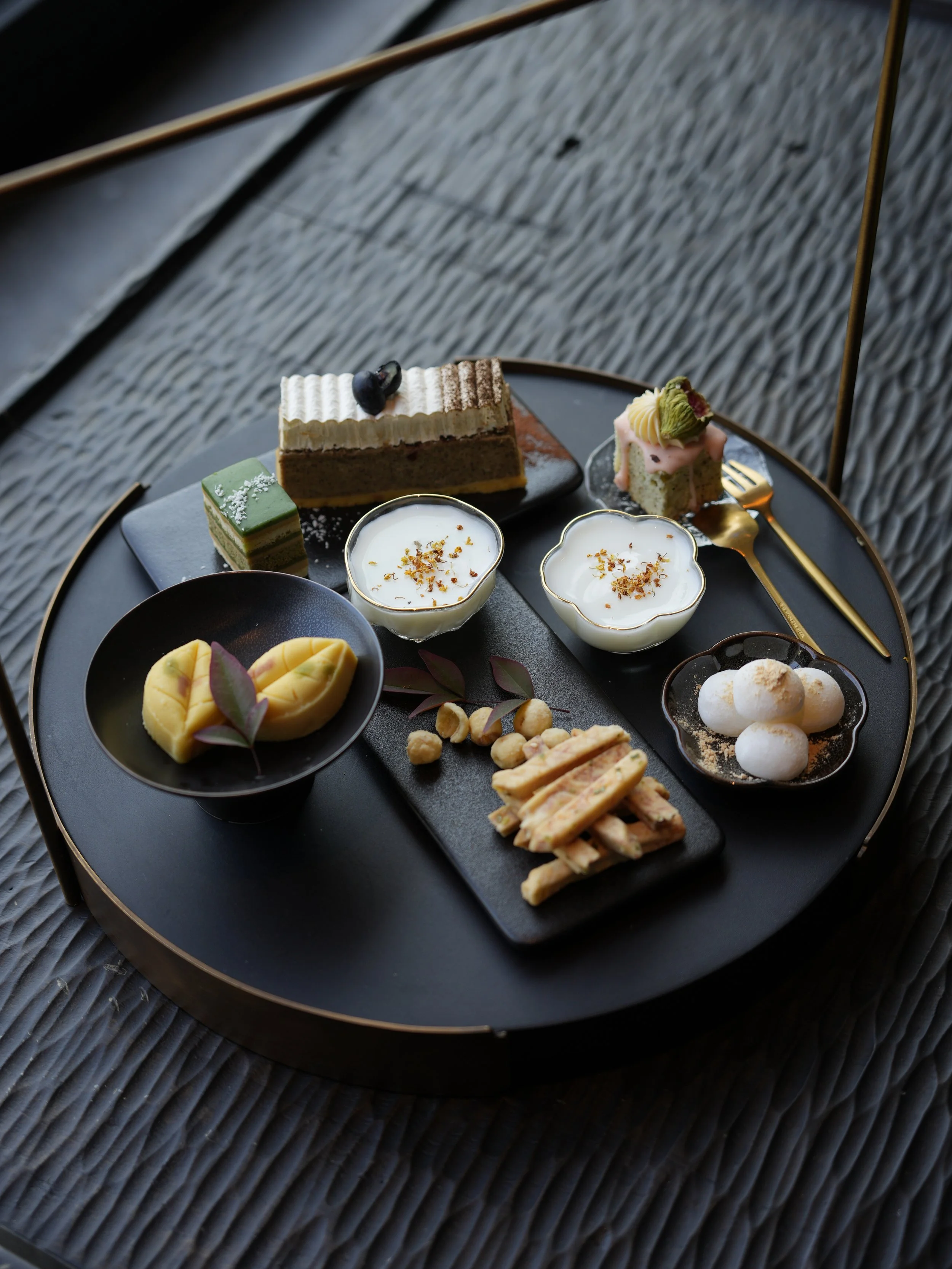 Assorted desserts on a black round tray, including a layered cake with blueberries, a green square cake with white sprinkles, two cups of white pudding topped with herbs and crumbs, a pink cake with cream and a green topping, a bowl of peeled mango s
