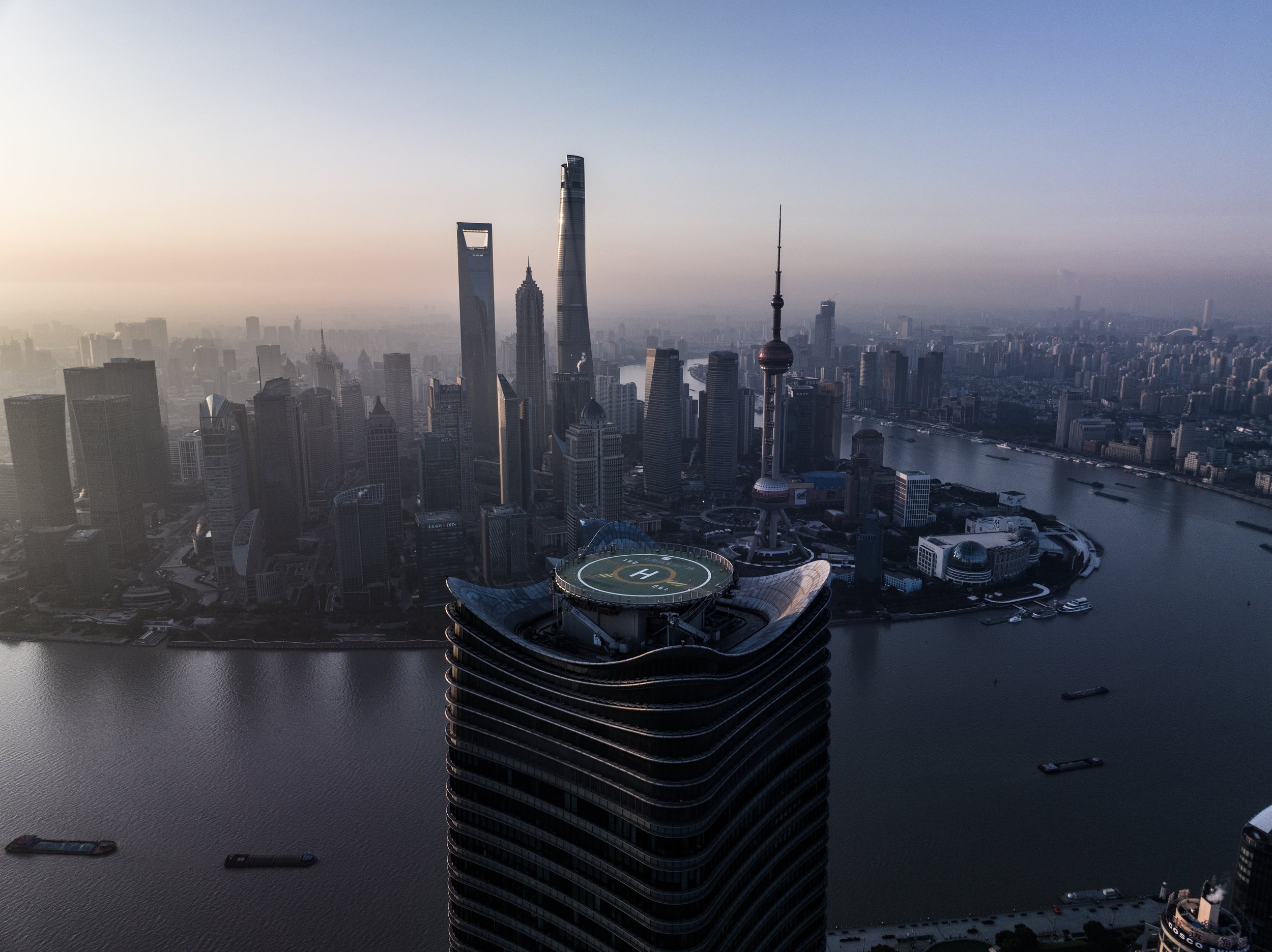 A city skyline featuring several skyscrapers, including the Shanghai Tower, Shanghai World Financial Center, and Jin Mao Tower, with a river in the foreground and a helipad on a nearby building.