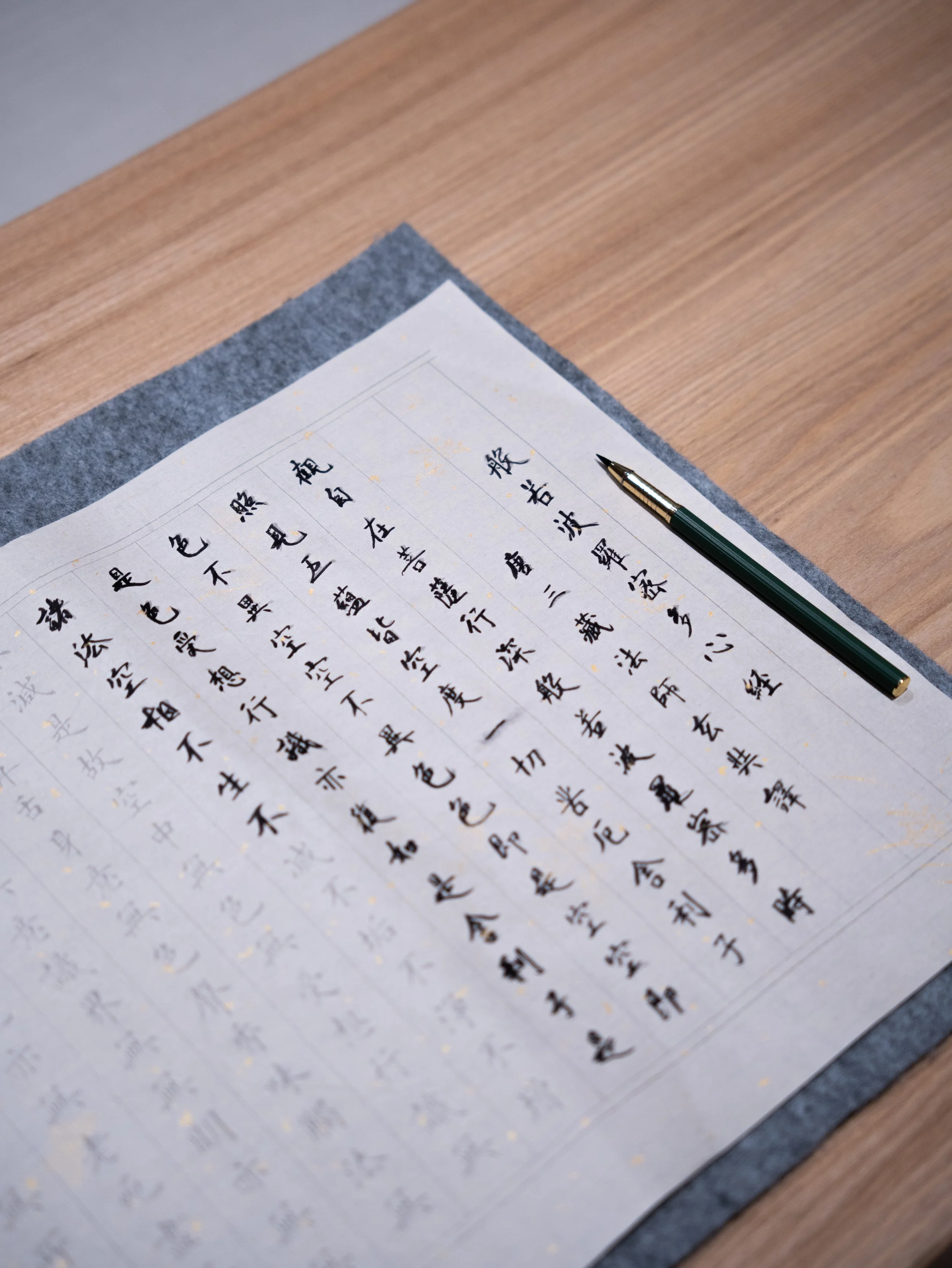 A sheet of paper with Chinese calligraphy characters written in black ink, placed on a gray mat on a wooden surface, with a green and gold pen resting on the paper.