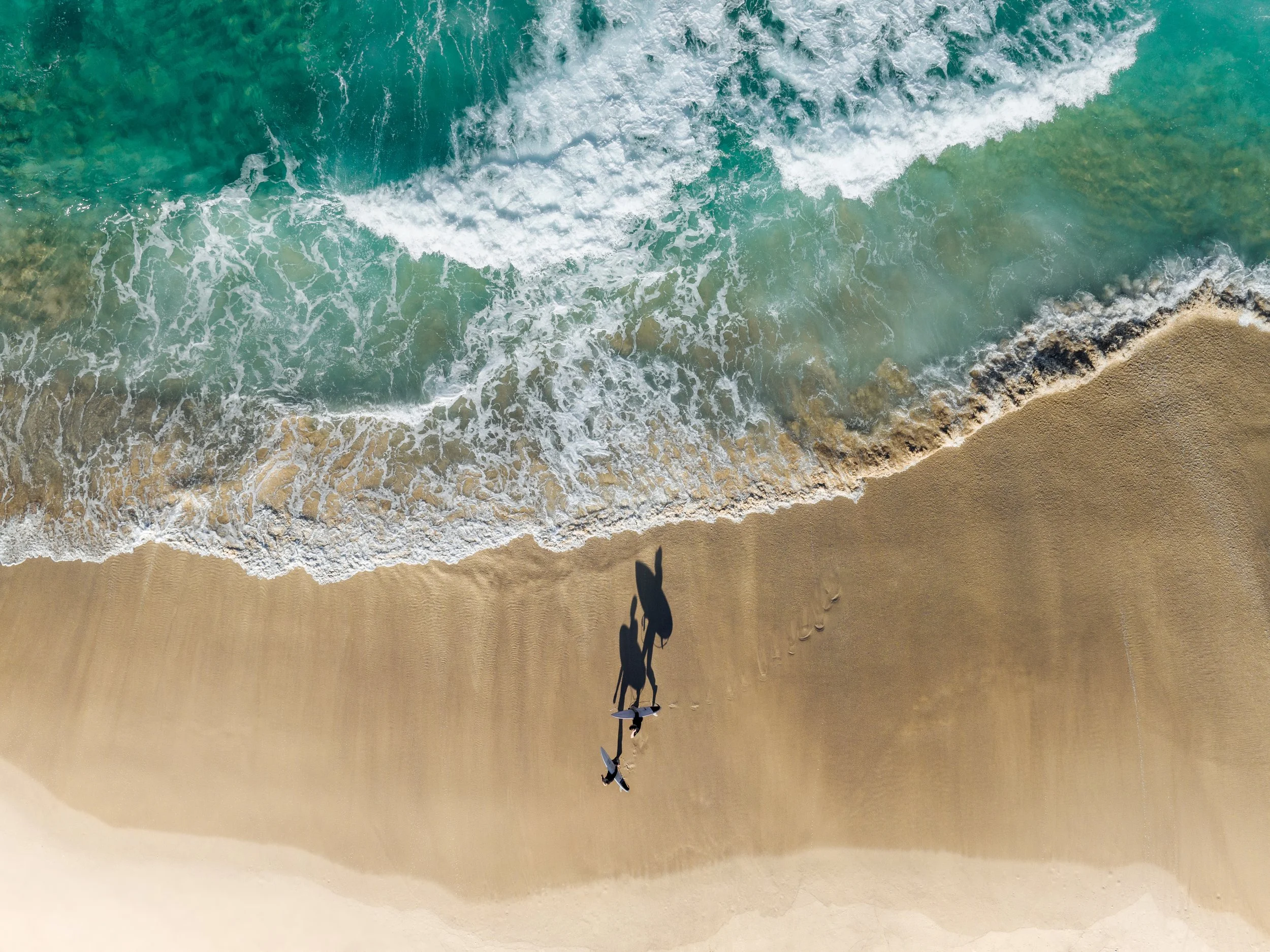 A shadow of a person riding a surfboard on the sandy beach, captured from above with waves approaching the shoreline.