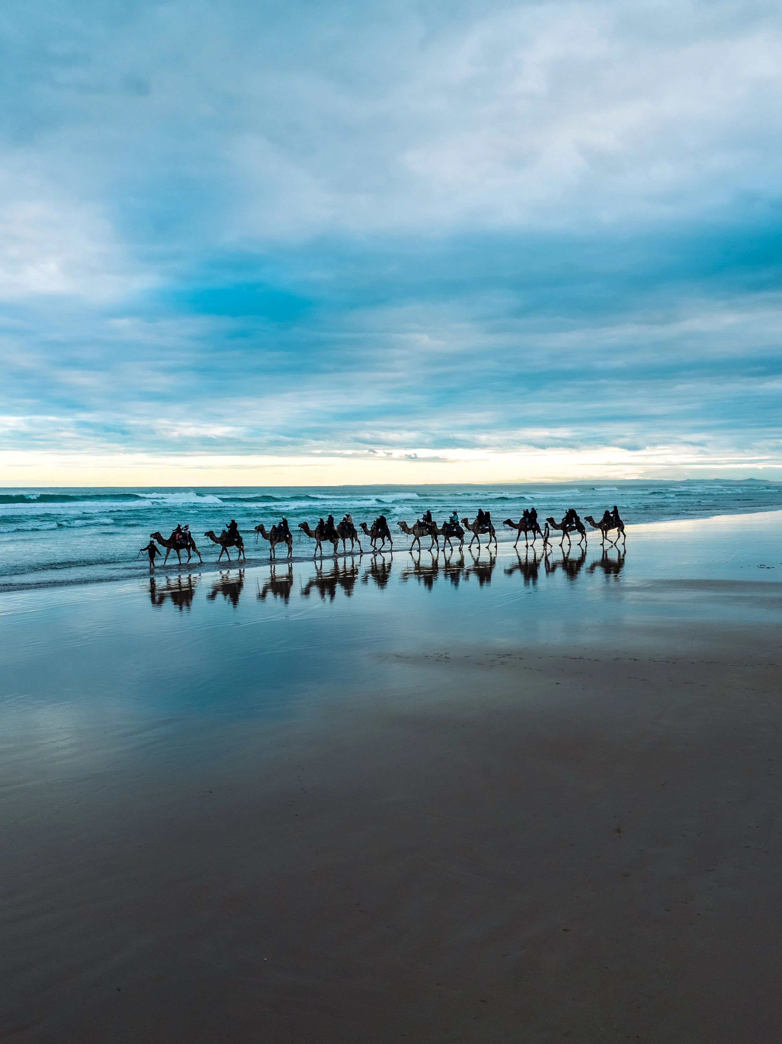 A line of people riding camels along the beach at sunset or sunrise, with clouds in the sky and reflections on wet sand.