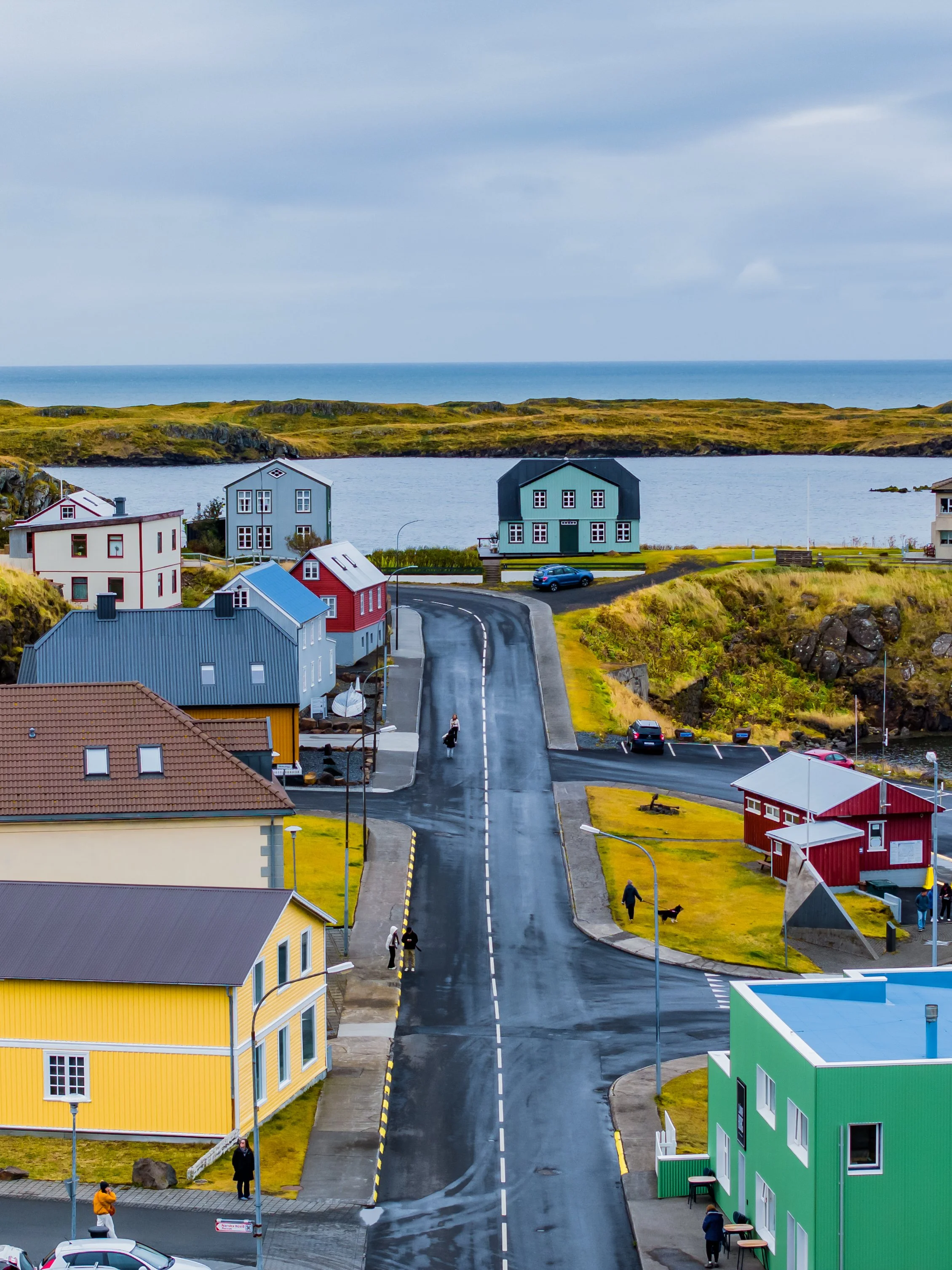 A street in a small town with colorful houses, including a yellow house, a green house, and a red house, with a body of water and land in the background under cloudy skies.