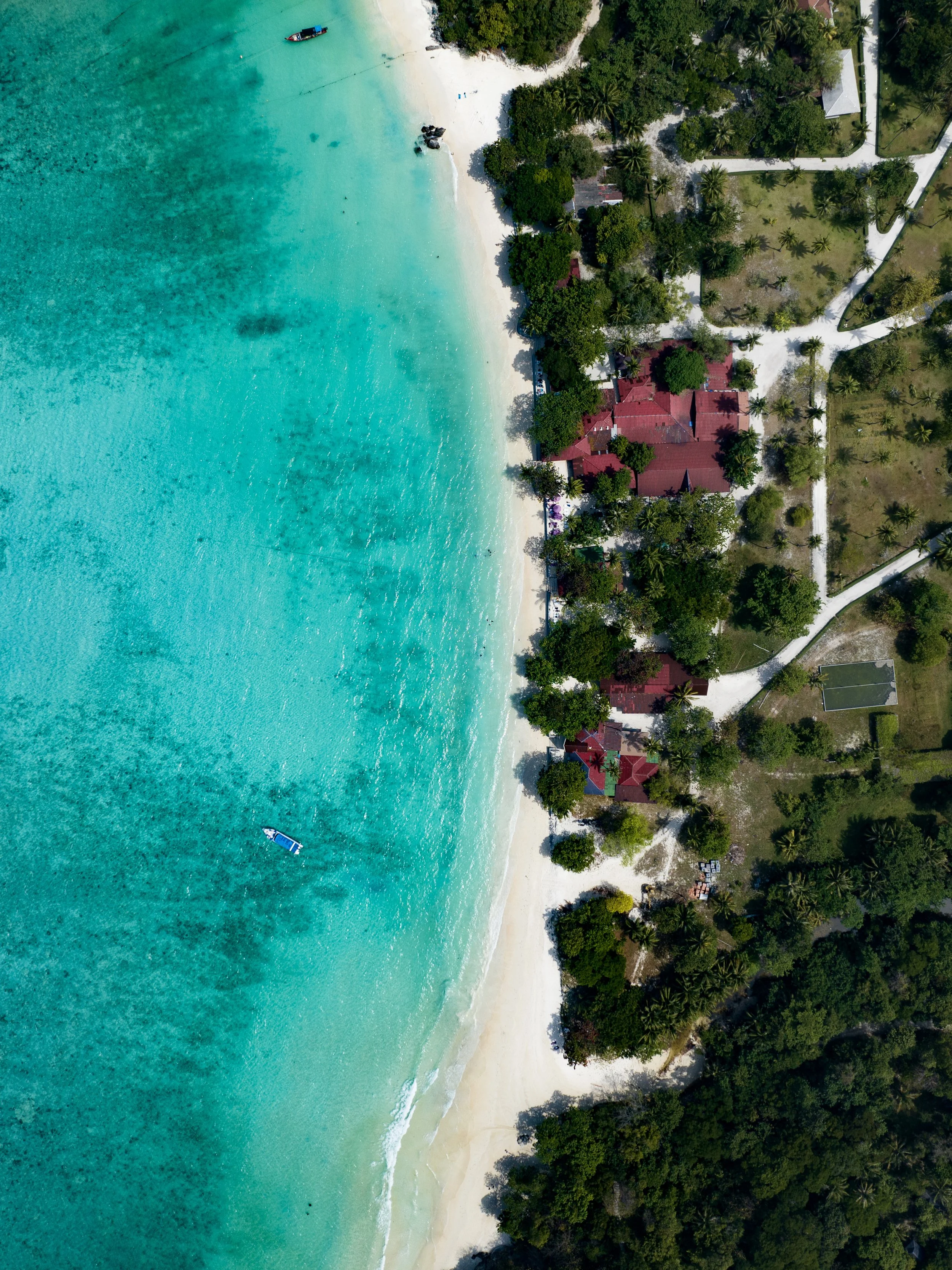 Aerial view of a tropical beach with clear turquoise water, white sandy shoreline, lush green trees, and red-roofed buildings along the coast.