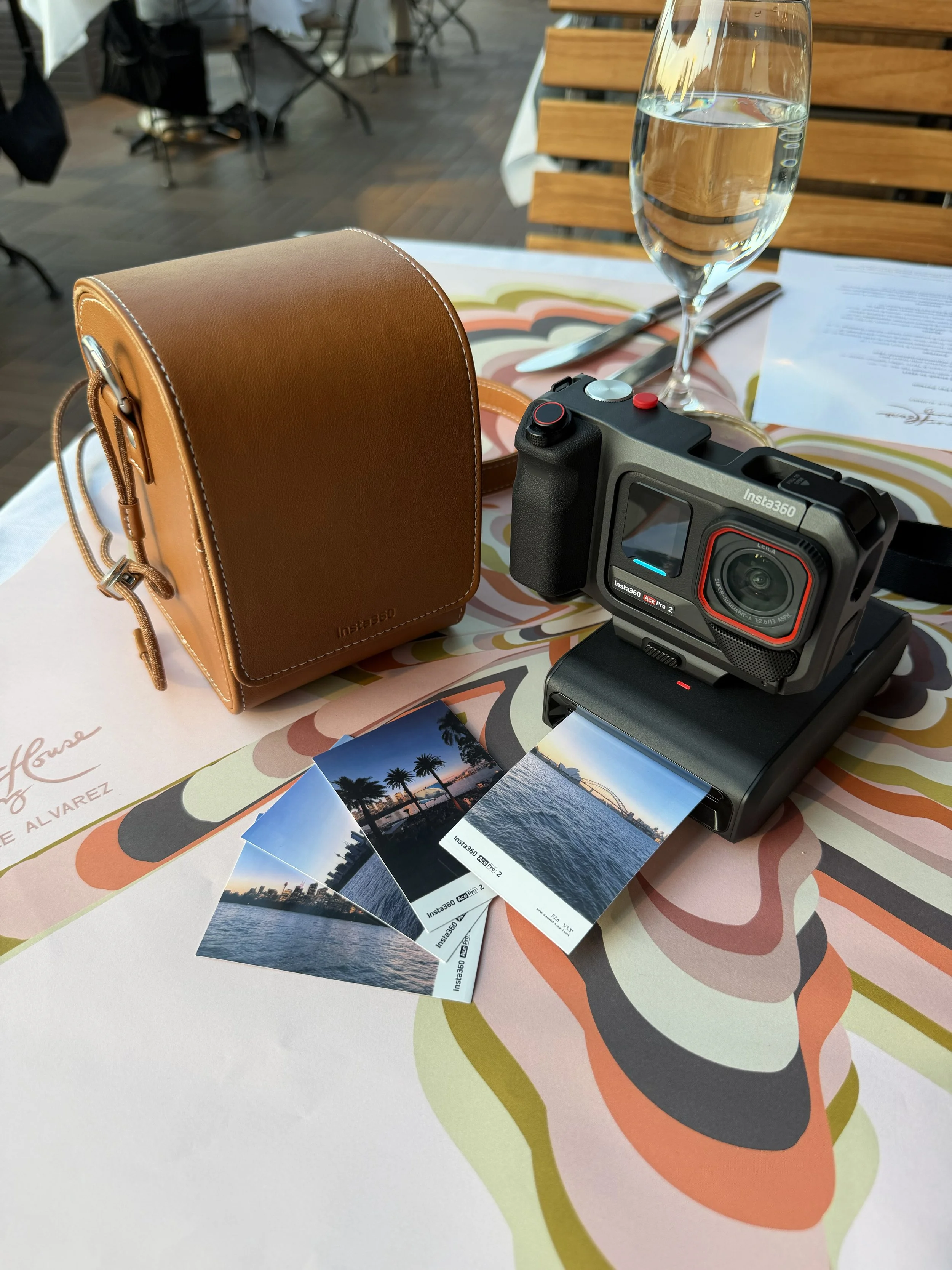 A tabletop in a restaurant with a leather bag, a camera, several printed photographs of a bridge and water, a glass of water, a knife, a fork, and a menu. The background shows wooden chairs and tables.