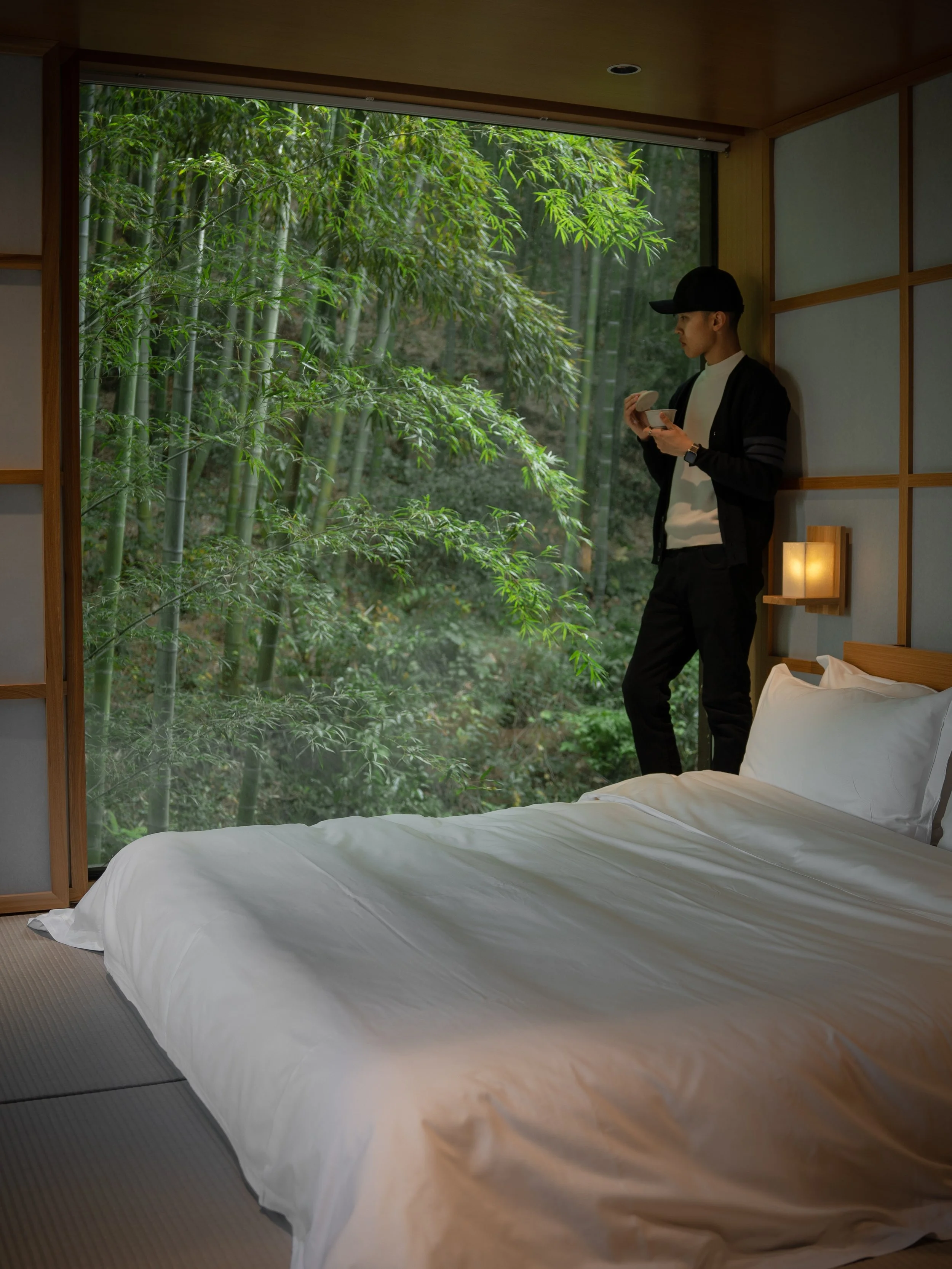 A person standing inside a room near a large window, looking outside at green bamboo trees.