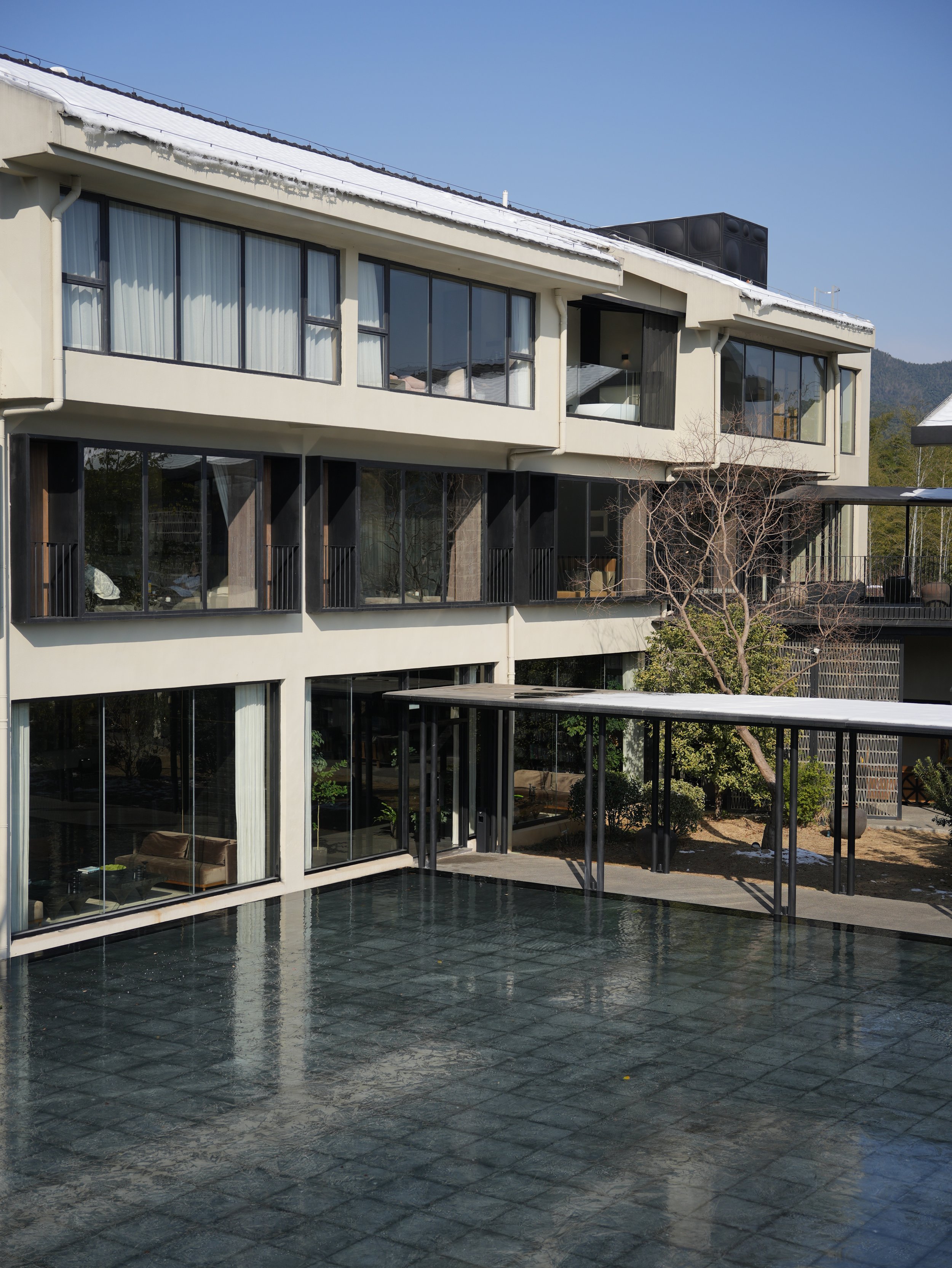 Modern multi-story building with large glass windows, a rooftop with snow, and a reflective pool in the foreground. Roof includes a black HVAC system, with trees and hills in the background.