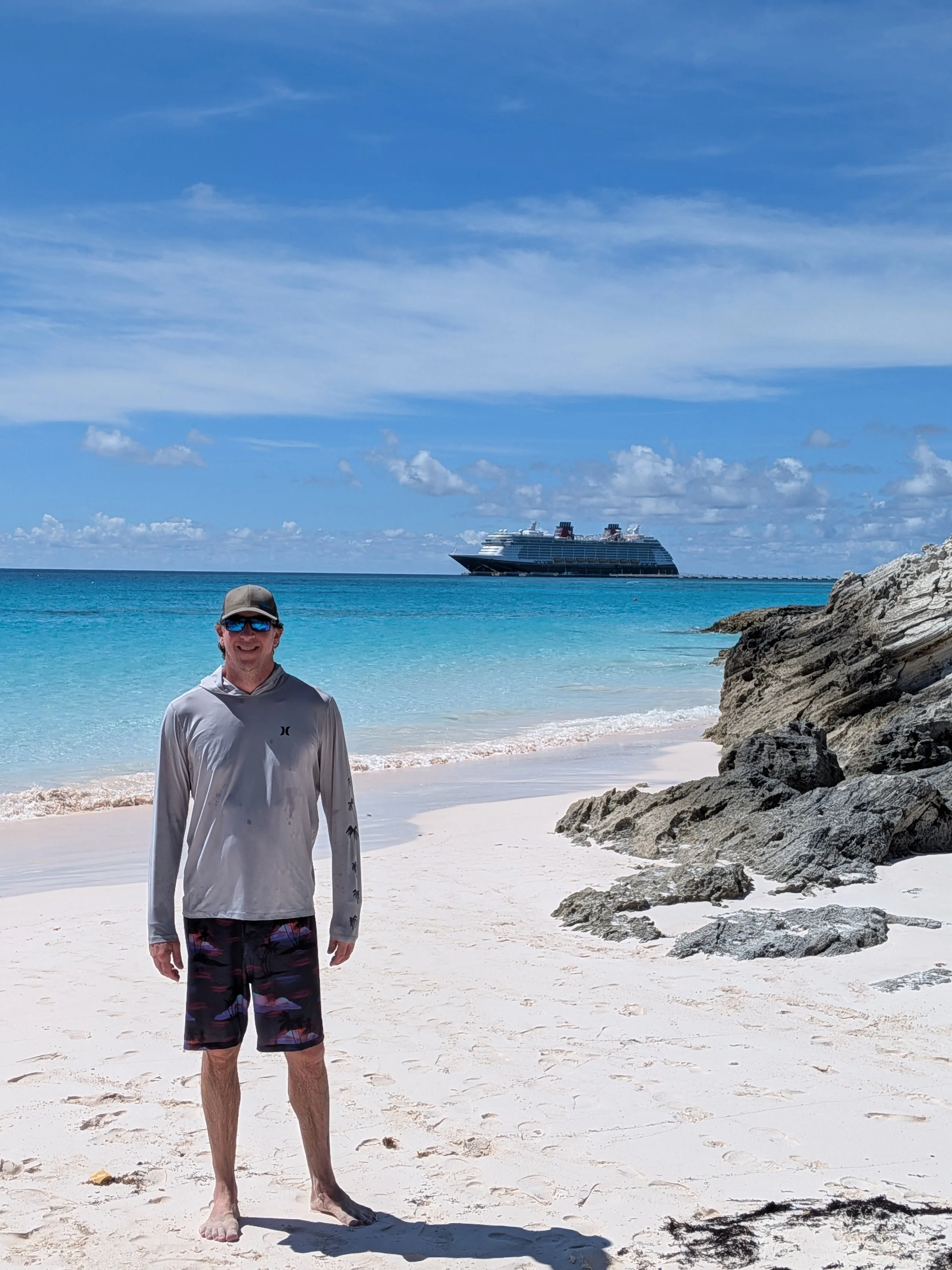 A man wearing sunglasses, a hat, a long-sleeve shirt, and shorts standing on a sandy beach with clear turquoise water, rocks, a cruise ship in the distance, and partly cloudy blue sky.
