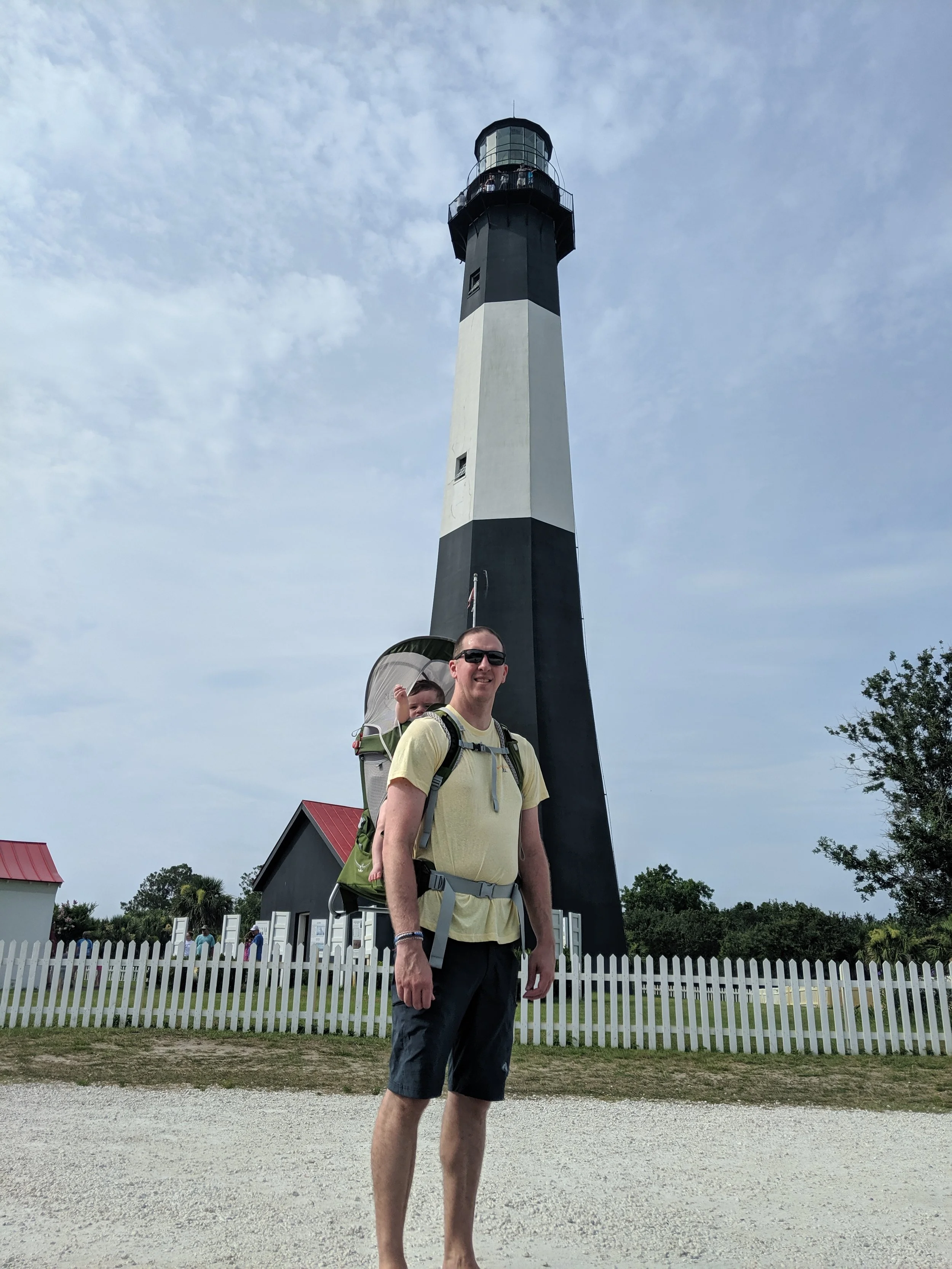 Man carrying a young child in a backpack carrier in front of a black and white lighthouse.
