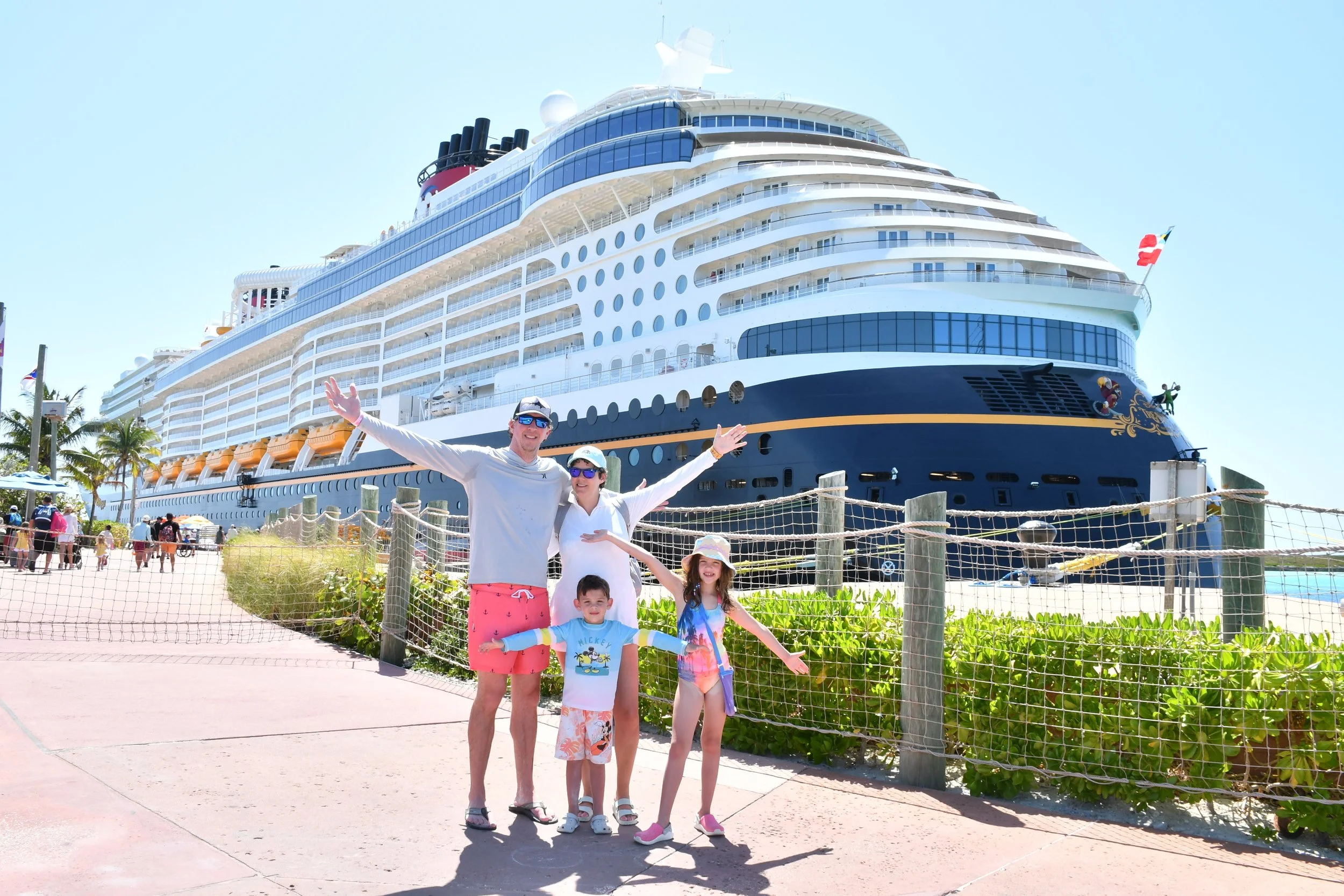 A group of four people, including two adults and two children, standing in front of a large cruise ship docked at a port on a sunny day. The group is smiling and waving, with the cruise ship visible in the background.