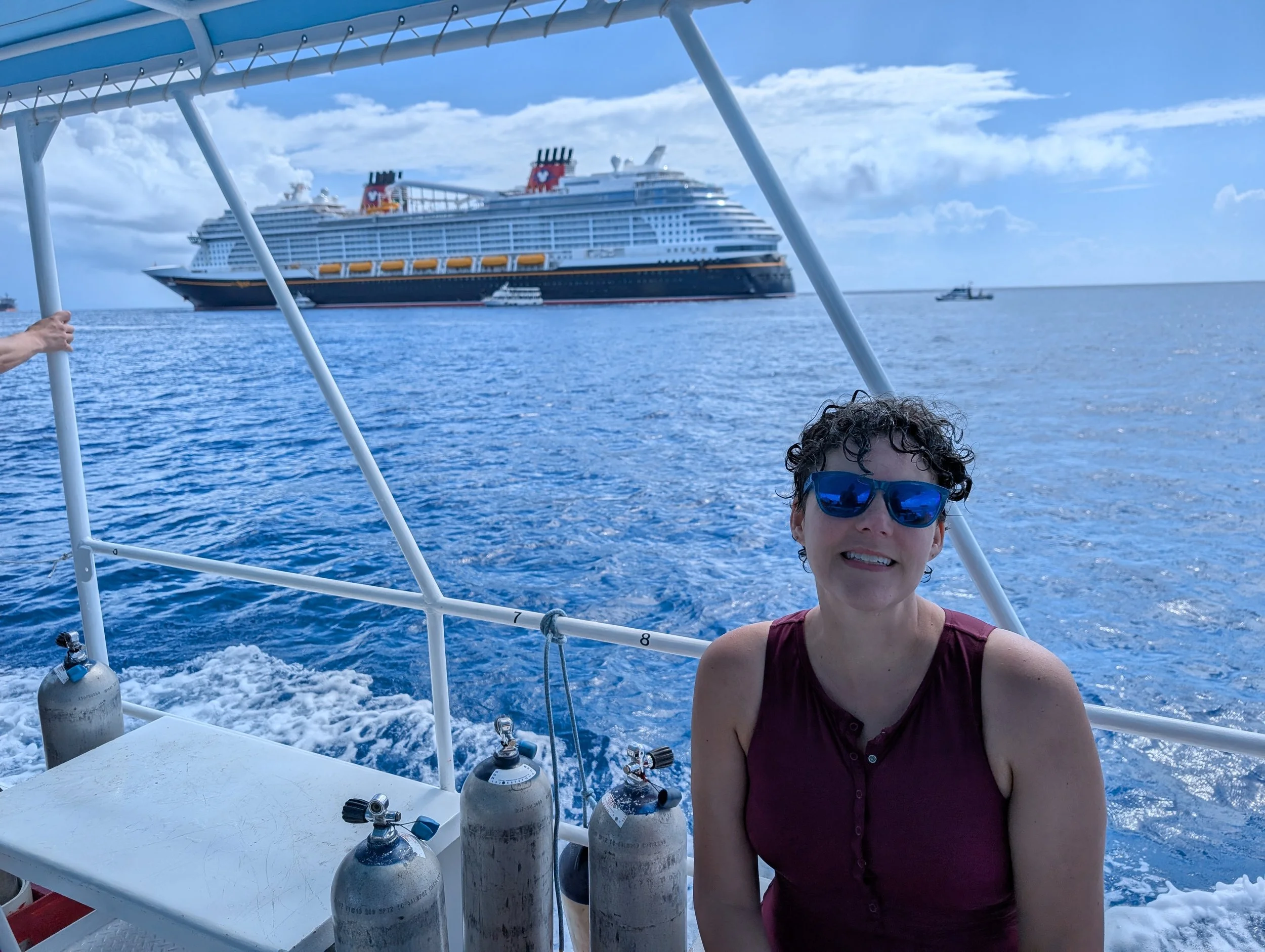 A woman wearing sunglasses on a boat with several scuba tanks in front. In the background, a large cruise ship with a black hull, red and yellow funnels, and a white superstructure is visible on the ocean under a partly cloudy sky.