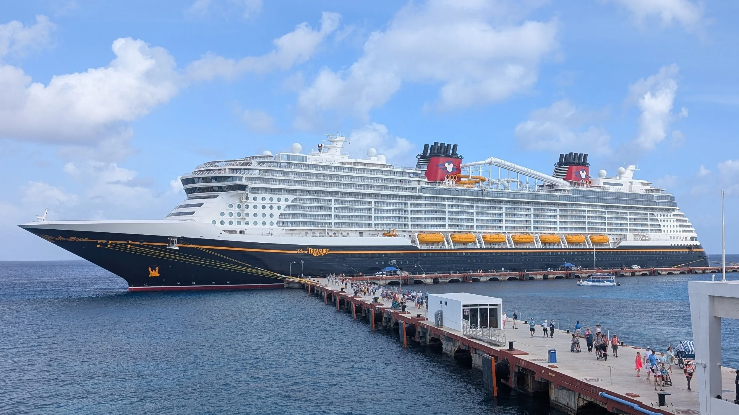 Disney Treasure cruise ship docked at a pier with passengers walking along the waterfront.