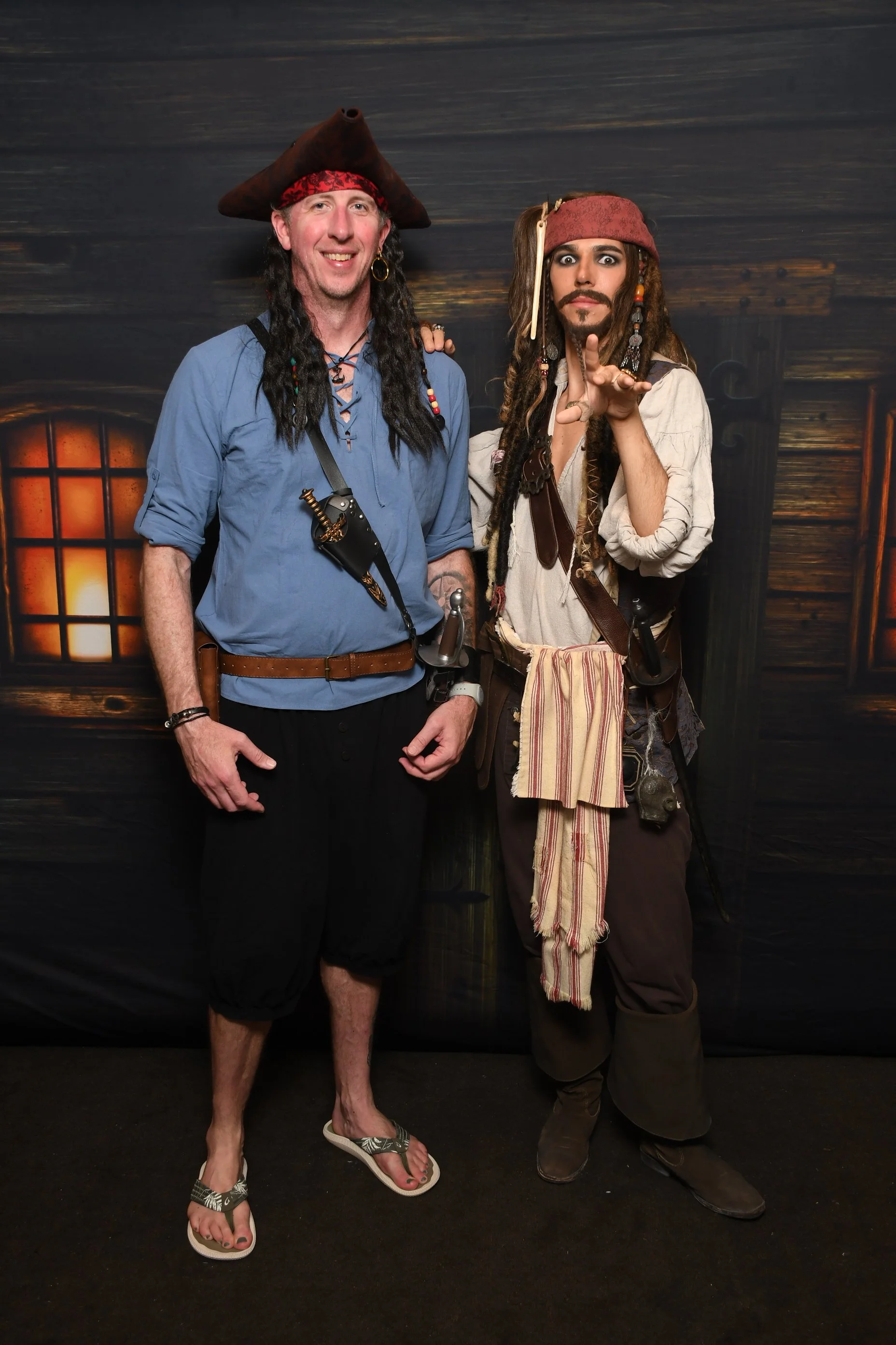 Two men dressed as pirates standing in front of a dark wooden background with glowing windows, one with traditional pirate attire including a hat and jewelry, the other with a more rugged costume.