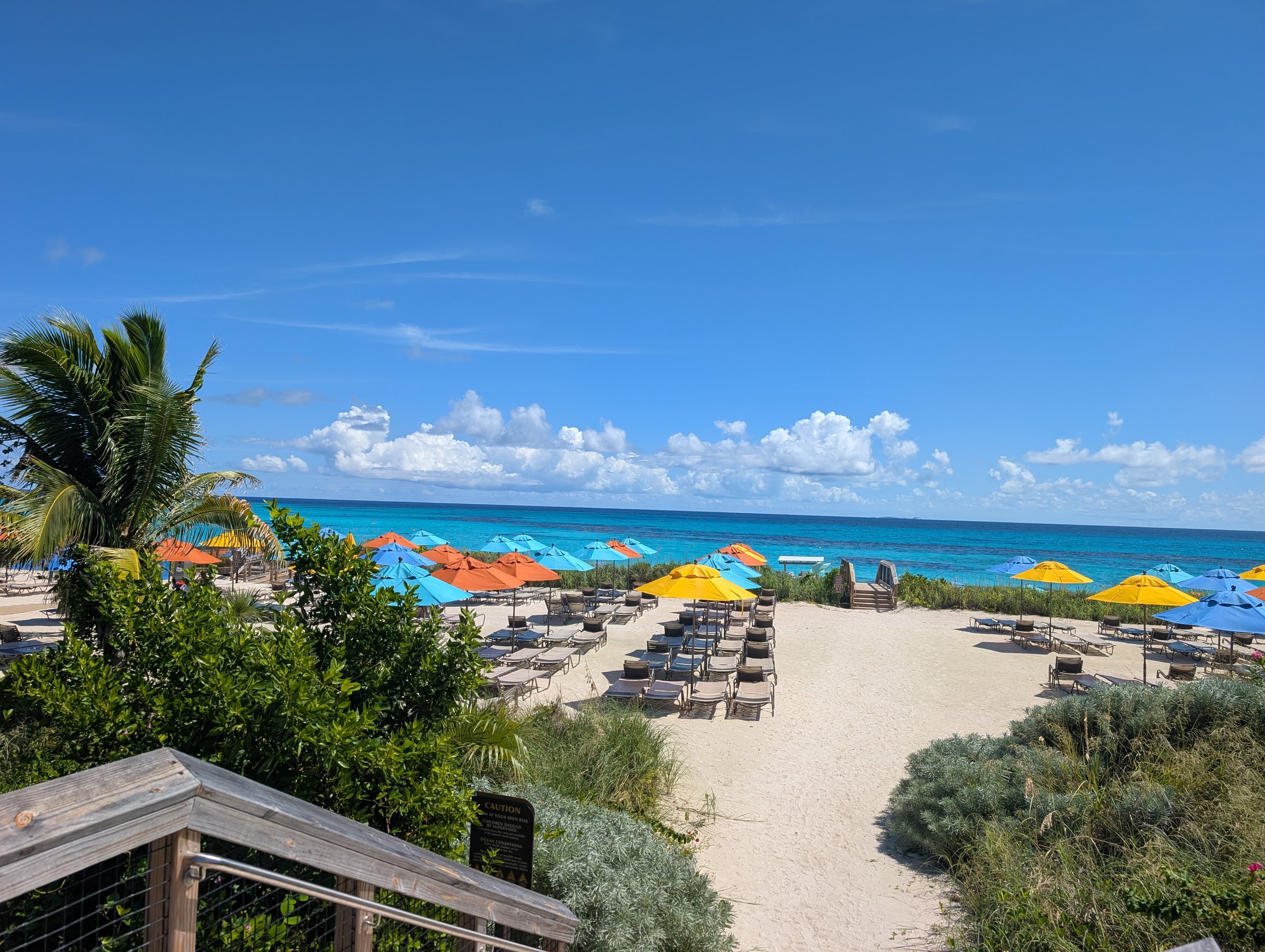 A beach with turquoise water, colorful umbrellas, lounge chairs, and palm trees under a mostly clear blue sky with some clouds.