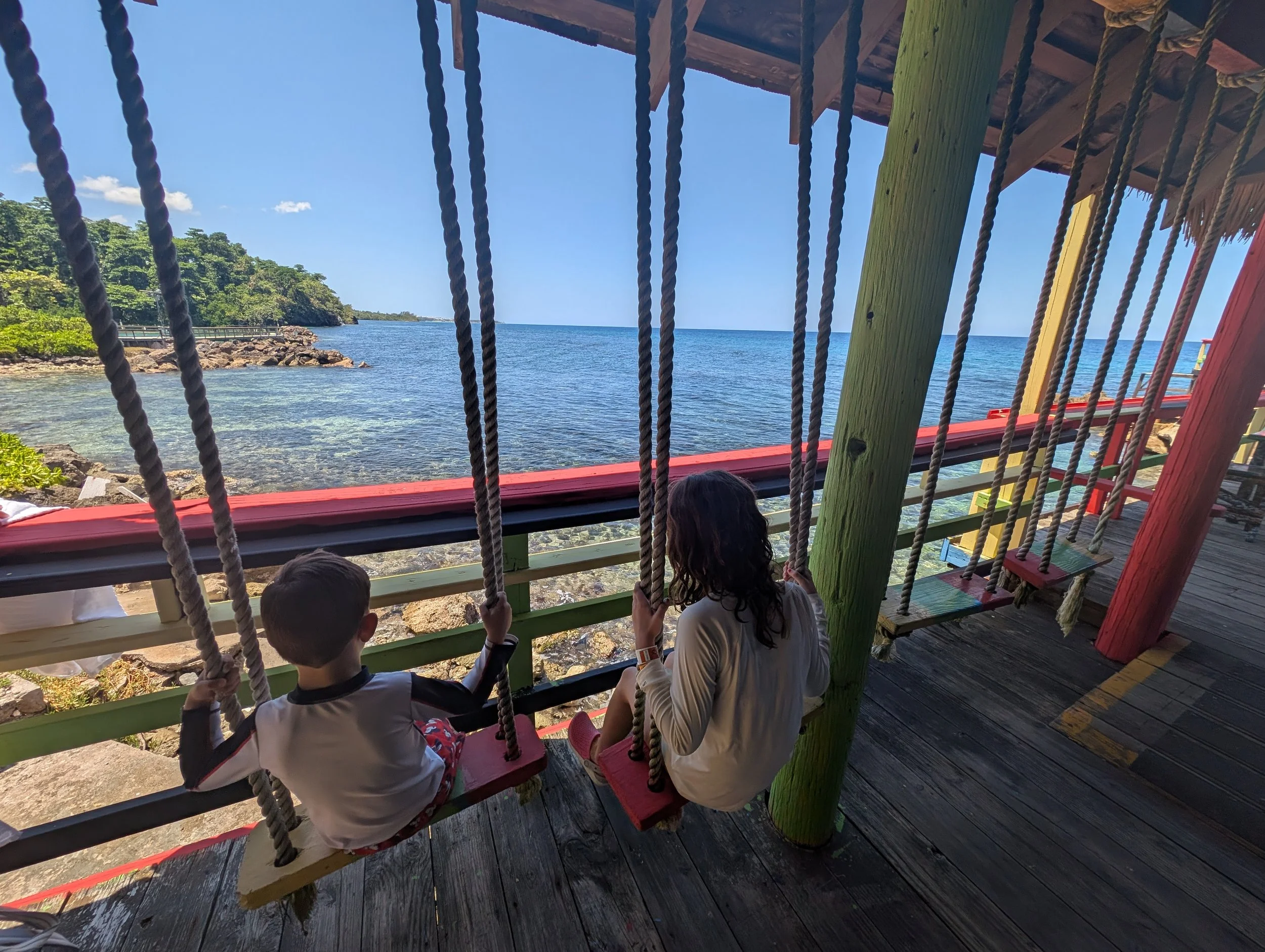 Children sitting on a colorful swing at a seaside park, with a view of the ocean, rocky shoreline, and green trees in the background, under a wooden shelter.