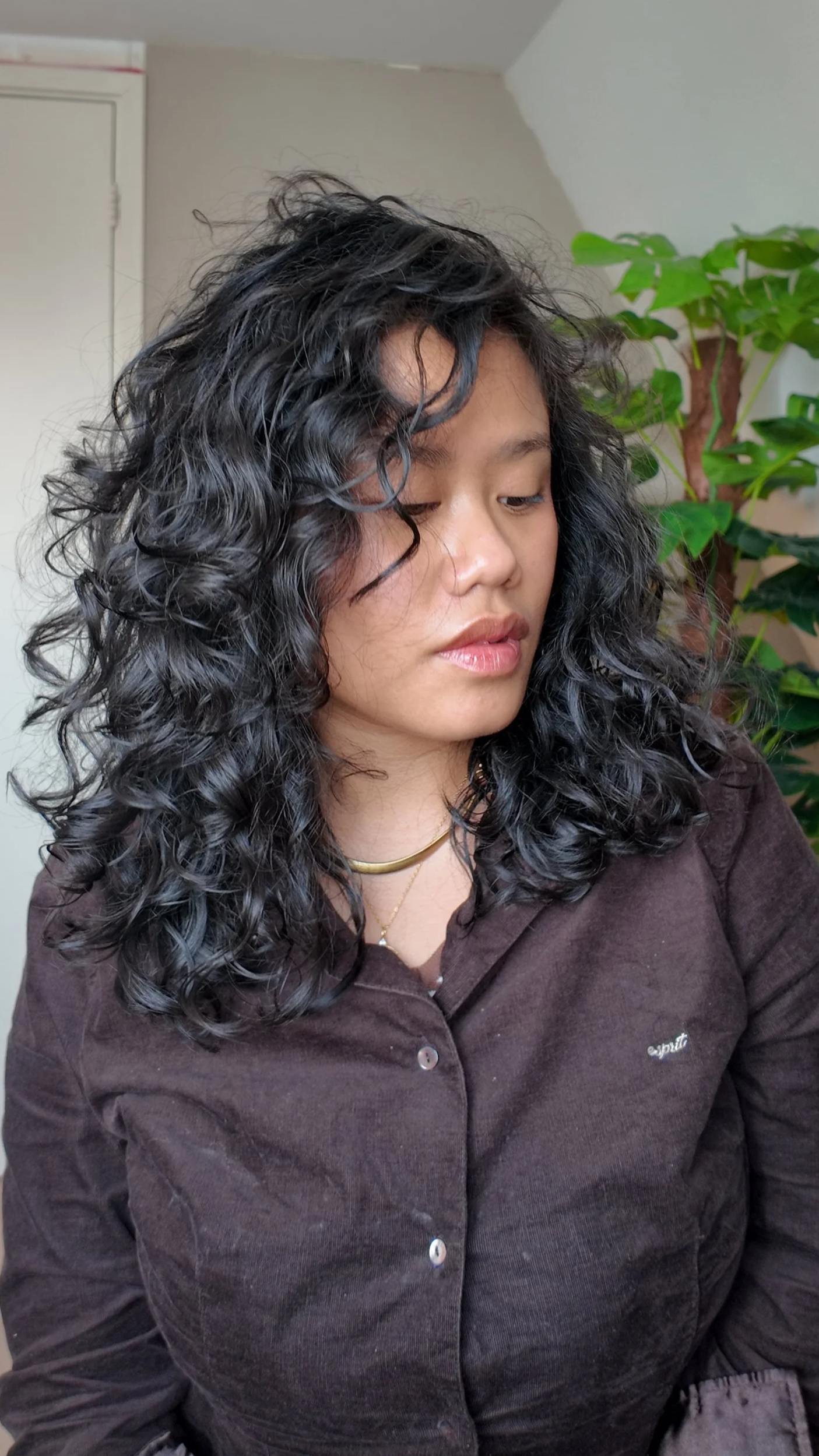 Close-up van een vrouw met krullend haar die naar beneden kijkt, in een binnenruimte met een plant op de achtergrond.