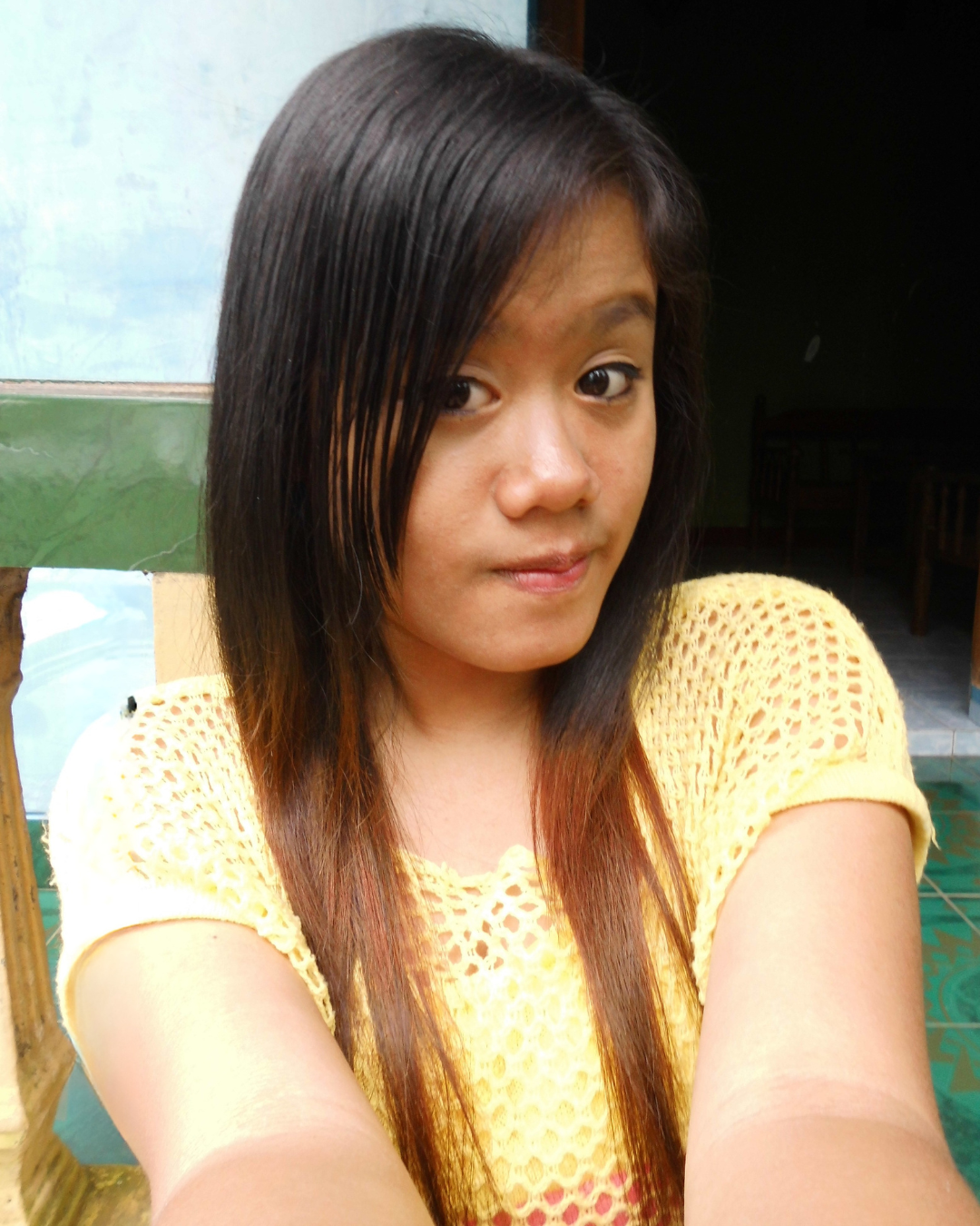 Young woman taking a selfie indoors, wearing a yellow knitted top, with straight black hair and a slight smile.