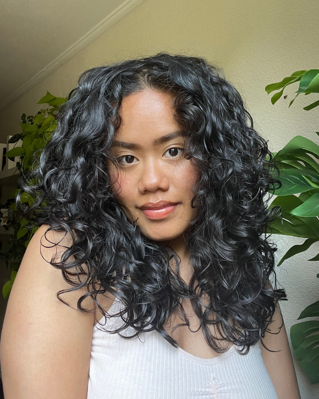 Een vrouw met lang, zwart, gekruld haar poseert binnenshuis met groene planten op de achtergrond.