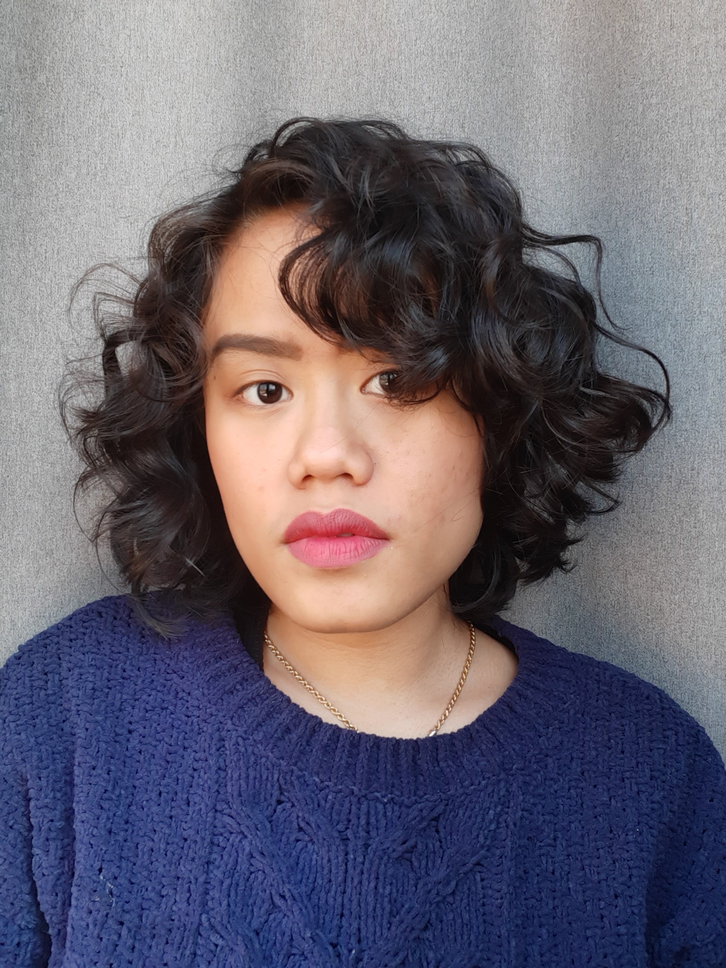 Close-up of a woman with curly black hair, wearing a dark blue textured sweater, a gold necklace, and a subtle makeup look with pink lipstick, against a plain gray background.