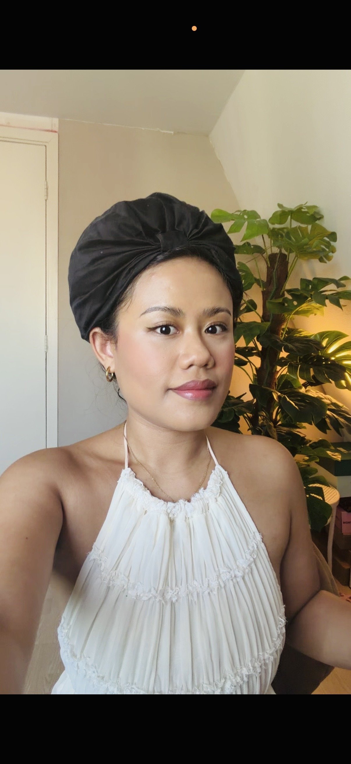 Vrouw met een zwarte haarband en een witte top poseert in een kamer met een groene plant op de achtergrond.