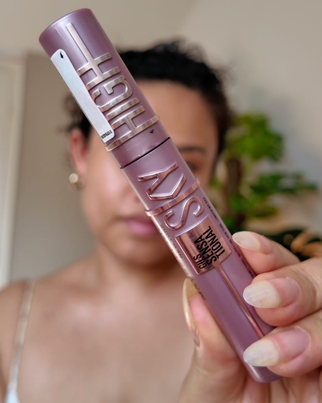 Close-up van een vrouw die een tube Maybelline The Falsies lash lift mascara vasthoudt, terwijl haar gezicht enigszins wazig op de achtergrond is.