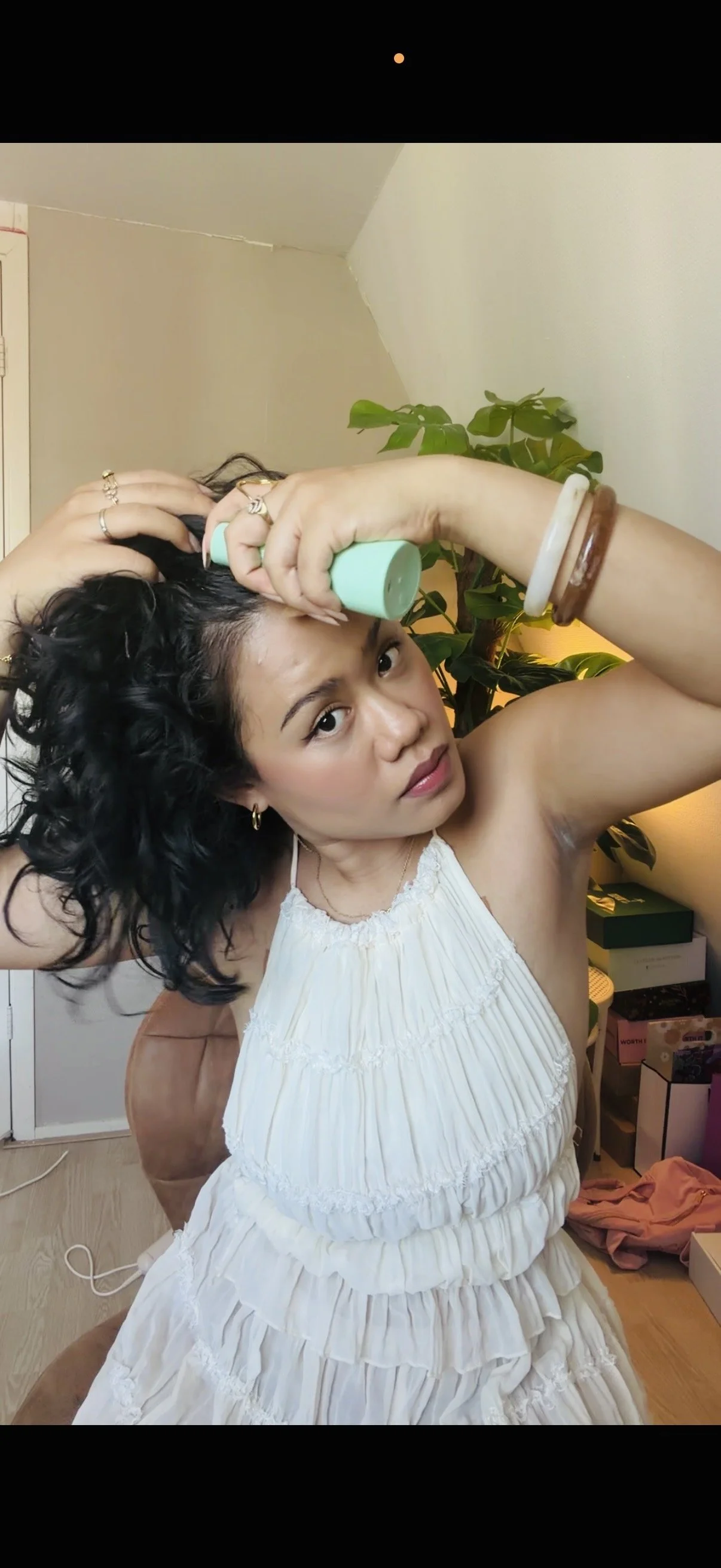 Vrouw met zwarte, krullende haren die haar haar kamt, terwijl ze een spray op haar haar spuit. Ze draagt een witte, ruffled jurk en zit op een bankje in een interieur met een groene plant op de achtergrond.
