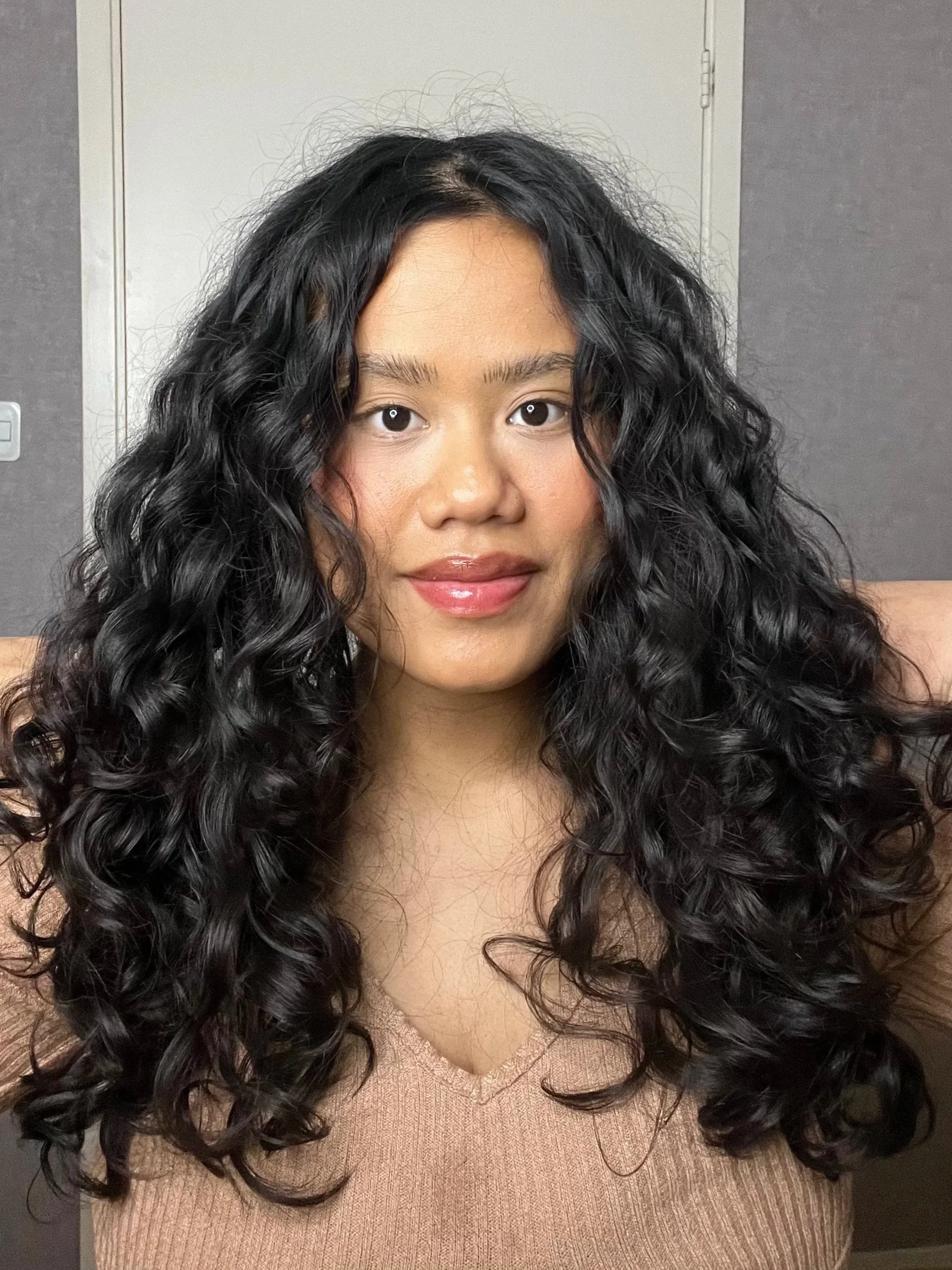 A woman with long, curly black hair wearing a beige V-neck sweater, smiling slightly, in front of a plain background.