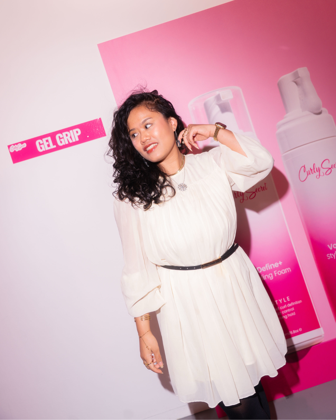 Vrouw poseert voor een roze en witte achtergrond met grote productverpakkingen, draagt een witte jurk met een zwarte riem, en wijst naar haar oor.