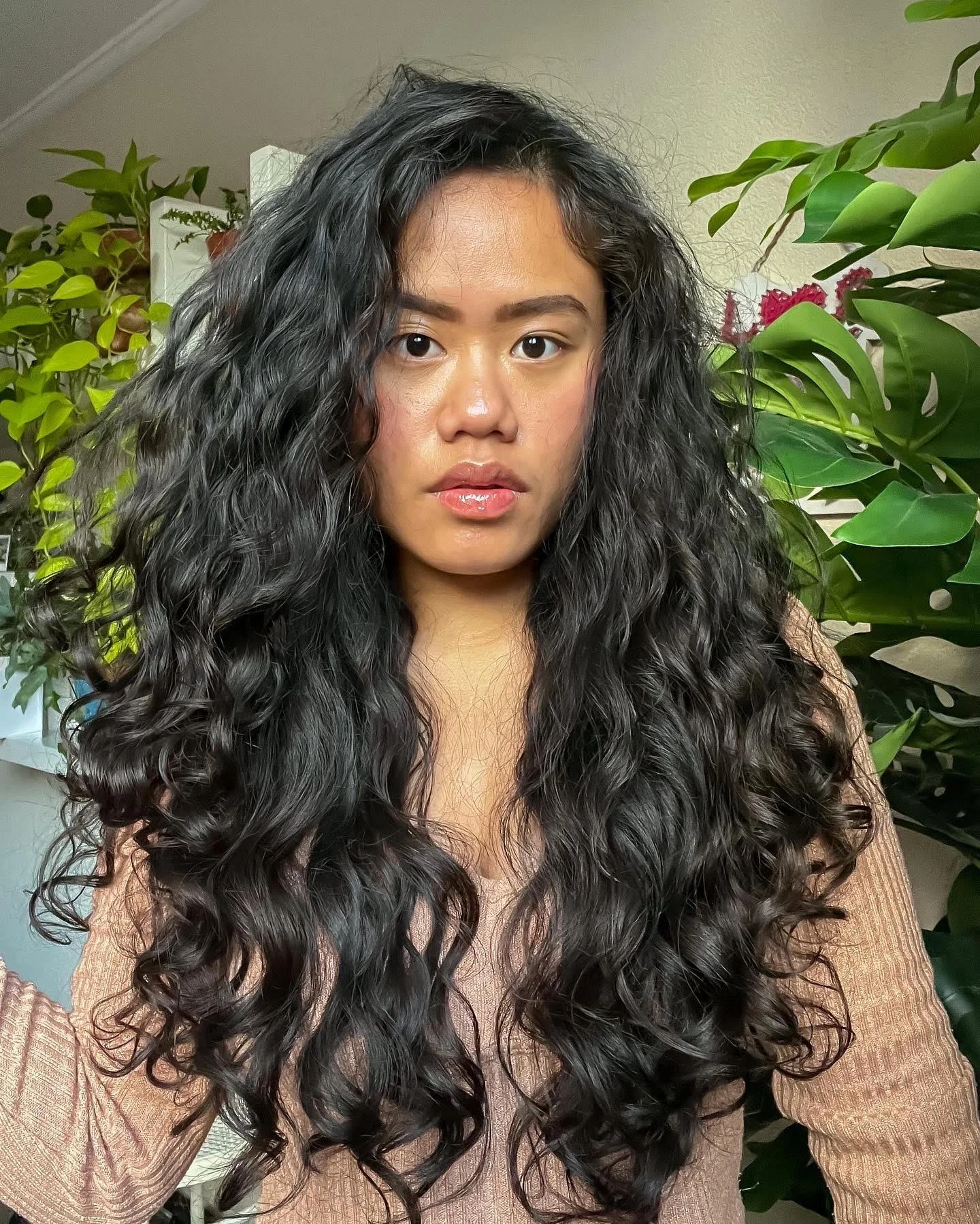 Close-up of a woman with long, curly black hair, wearing a beige sweater, standing in front of various green plants.