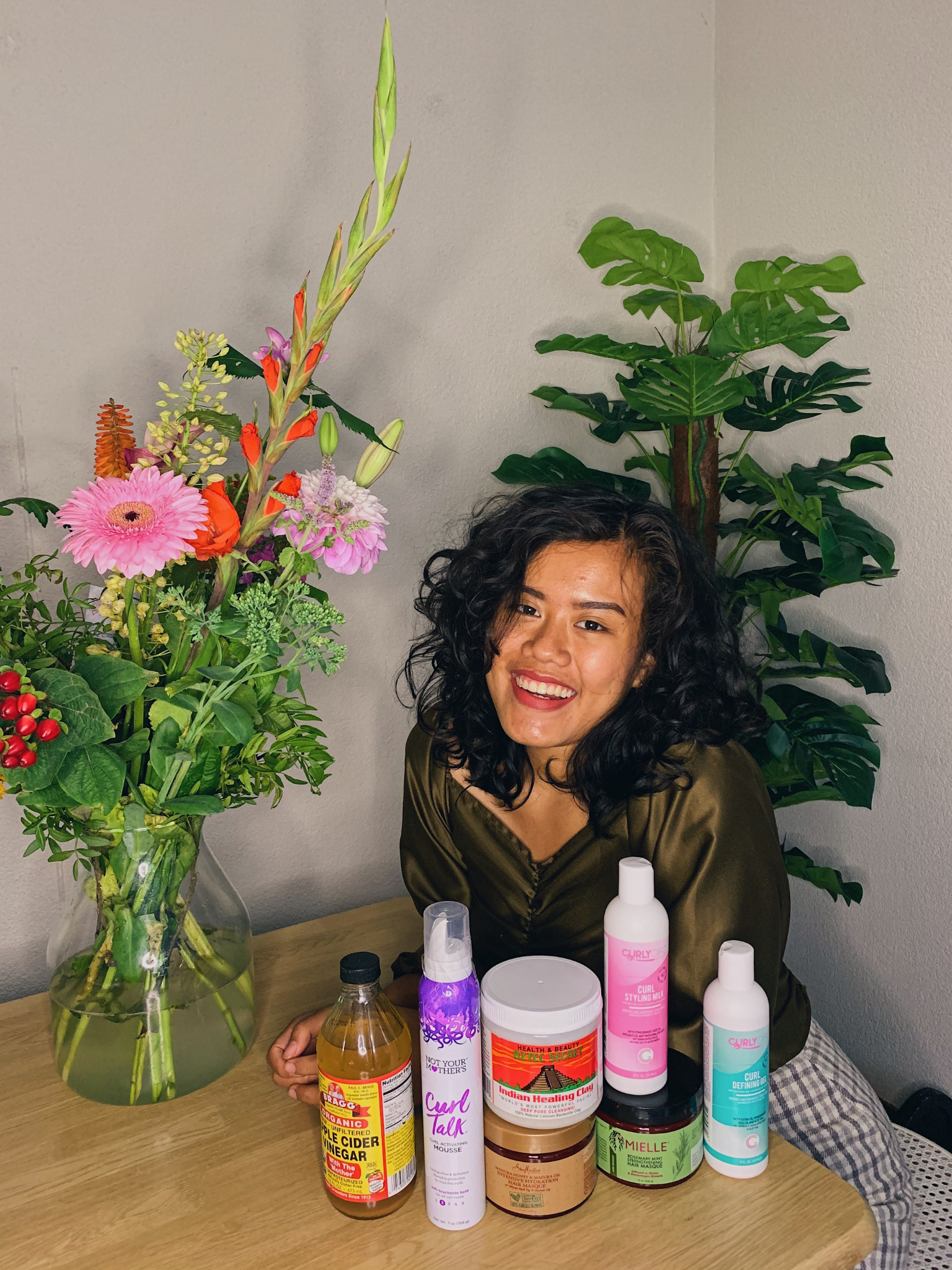A woman with curly black hair smiling at a table with various curly hair products, and a large floral arrangement in a glass vase, with green foliage and colorful flowers, behind her.