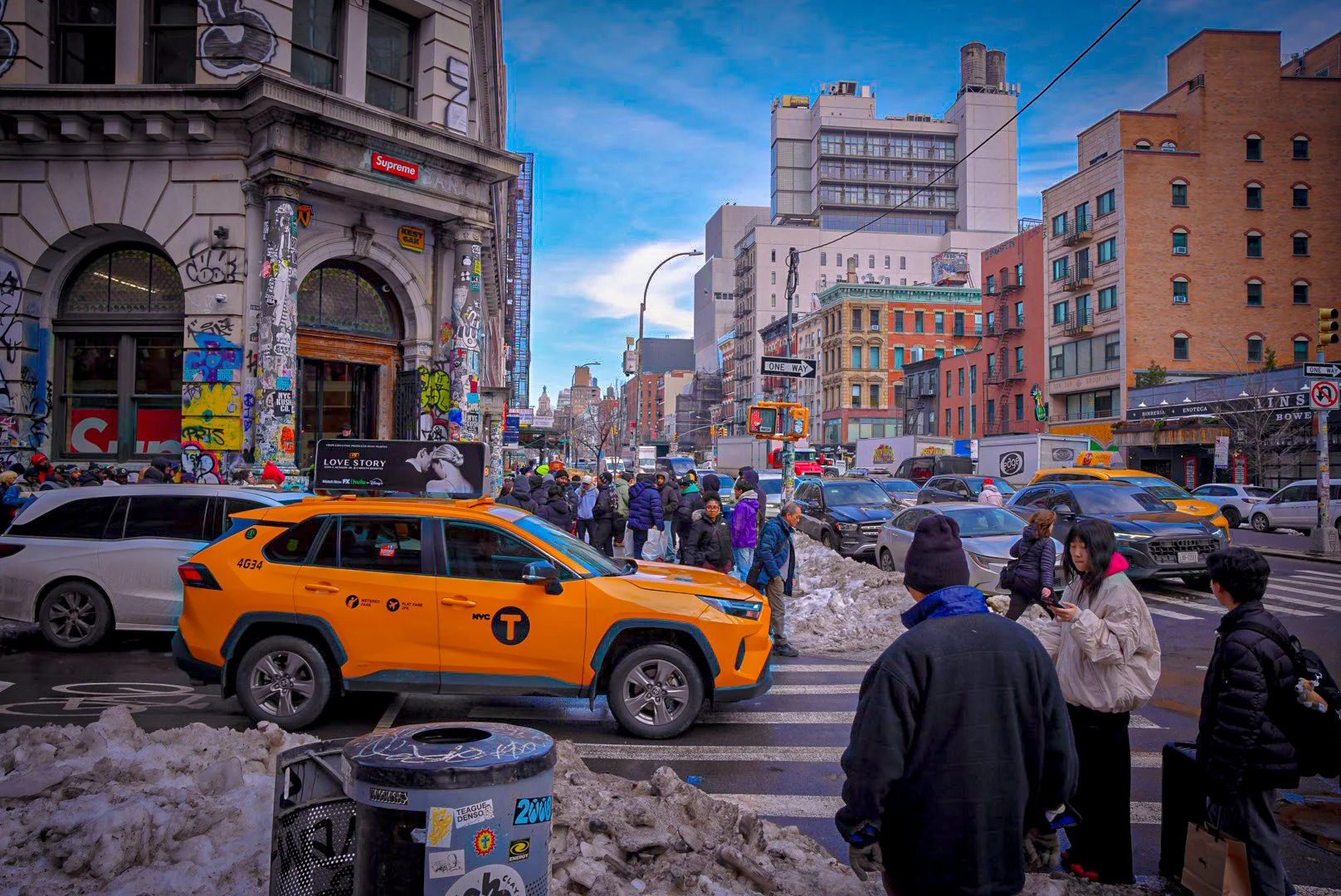 Street scene in New York City with pedestrians crossing at a crosswalk, yellow NYC taxis, snow piles, and tall buildings covered in graffiti and advertisements on a clear day.