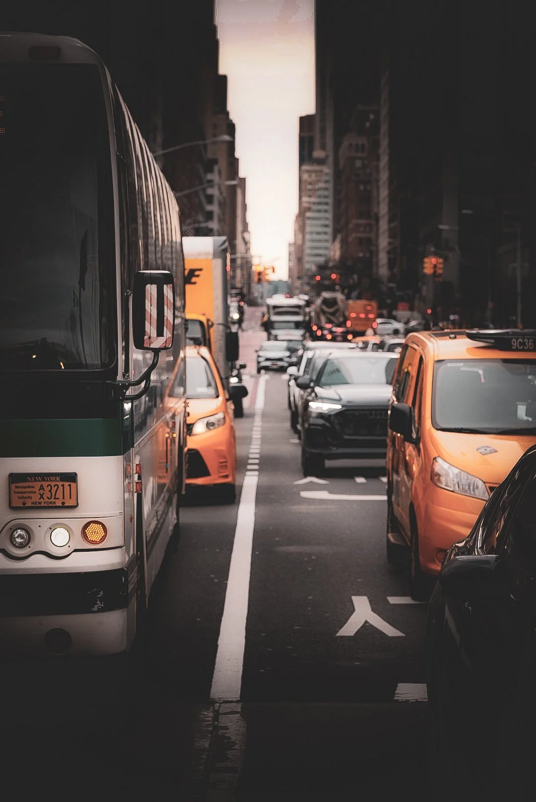 City street filled with cars, buses, and taxis during dusk with tall buildings on either side.