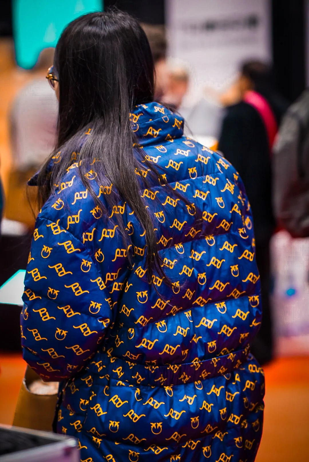 Back of a person wearing a blue jacket with a repeated pattern of yellow letters and symbols, standing in a crowded indoor space.