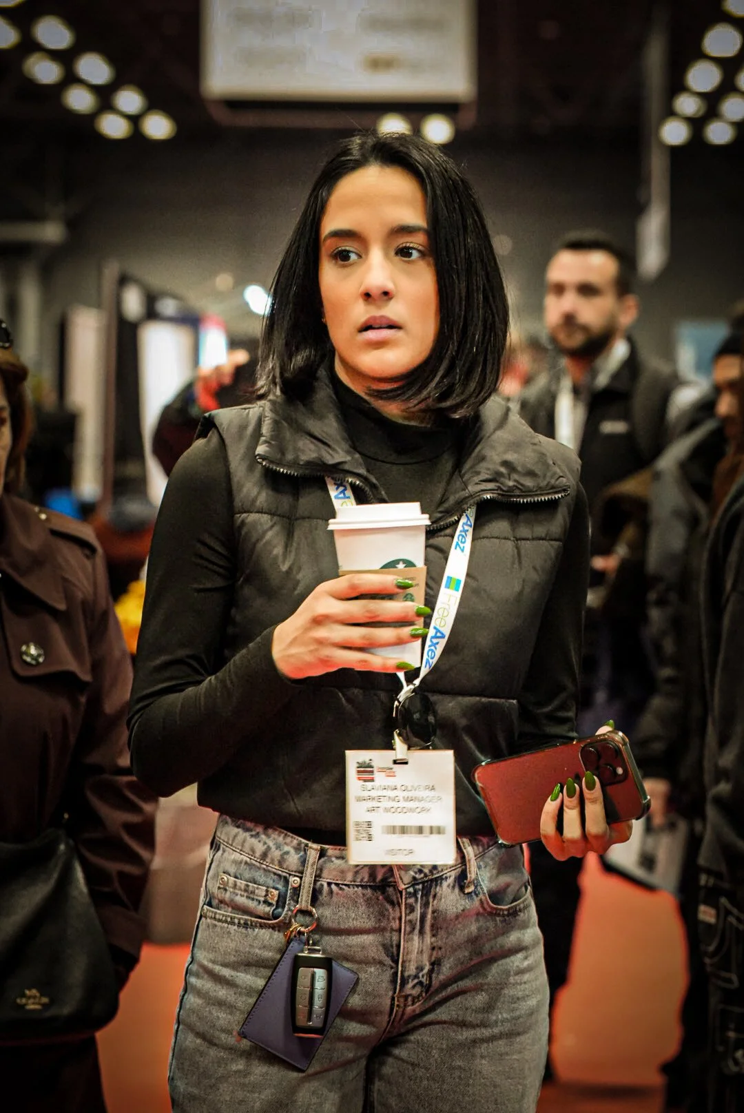 A woman with black hair holding a disposable coffee cup and a smartphone, wearing a black vest and jeans, standing in a busy indoor setting.
