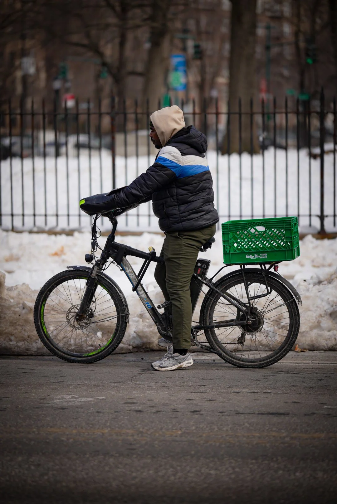 A man riding a black bicycle with a green crate attached to the back, wearing a puffy jacket, a hooded sweatshirt, and sneakers, on a street with snow piles and a metal fence in the background.