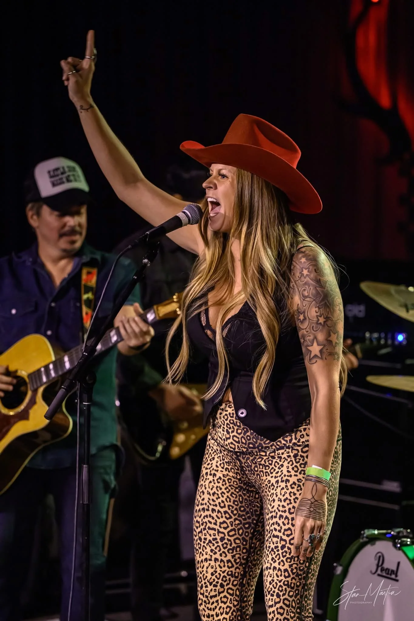 A woman with long wavy hair, tattoos on her left arm, wearing a red cowboy hat, black top, and animal print pants, singing into a microphone on stage. Someone in the background with a guitar, dark background with red lighting.