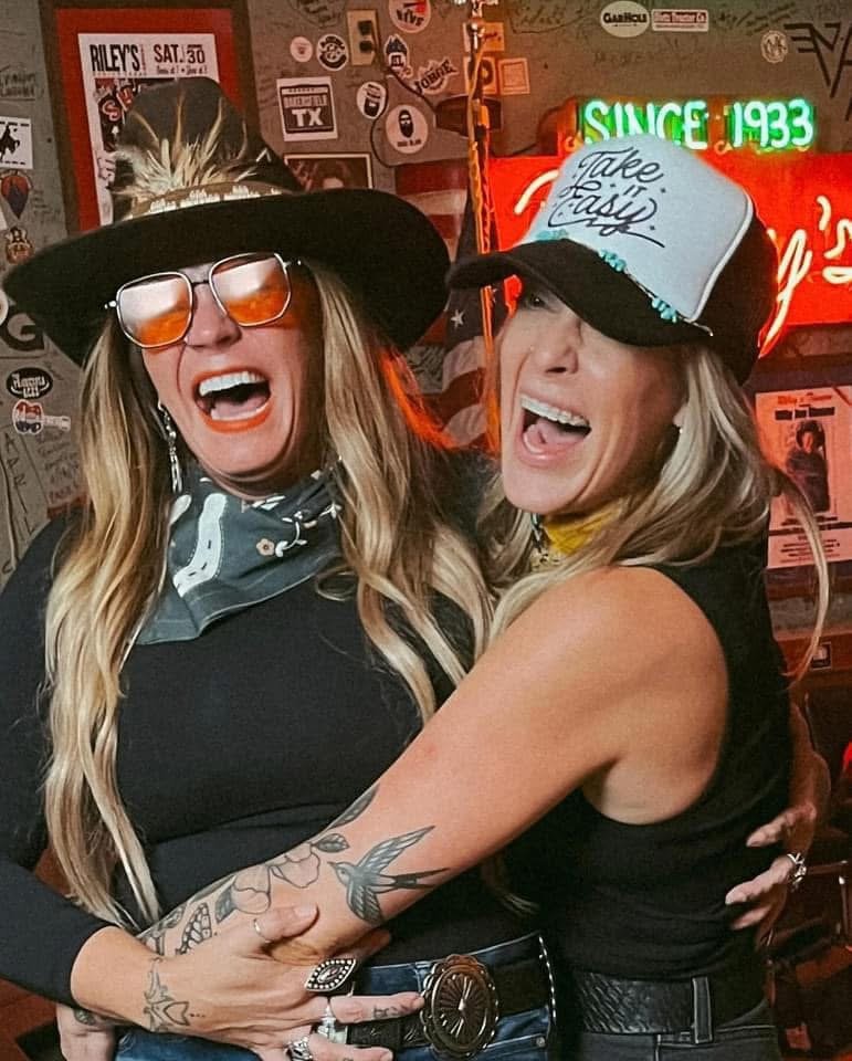 Two women in cowboy hats and sunglasses smiling and embracing each other in a lively bar decorated with neon signs and various posters.