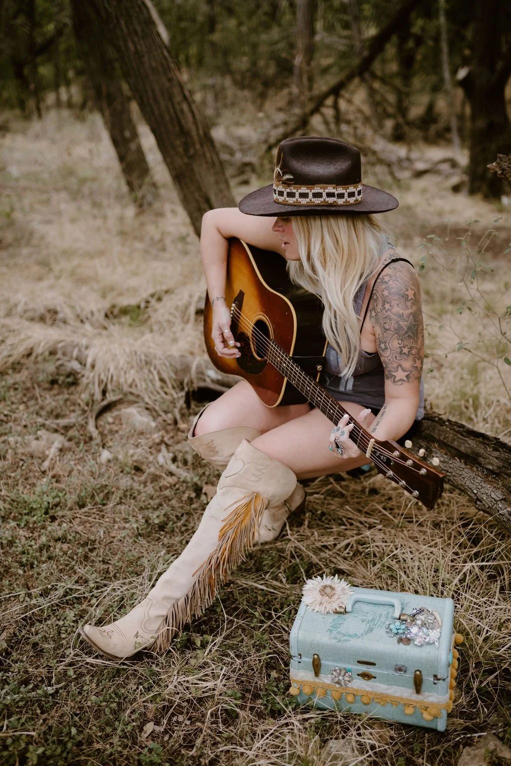 A woman sitting in a forest playing an acoustic guitar, wearing a wide-brimmed hat, cowboy boots, and a sleeveless top, with a decorated vintage jewelry box on the ground nearby.